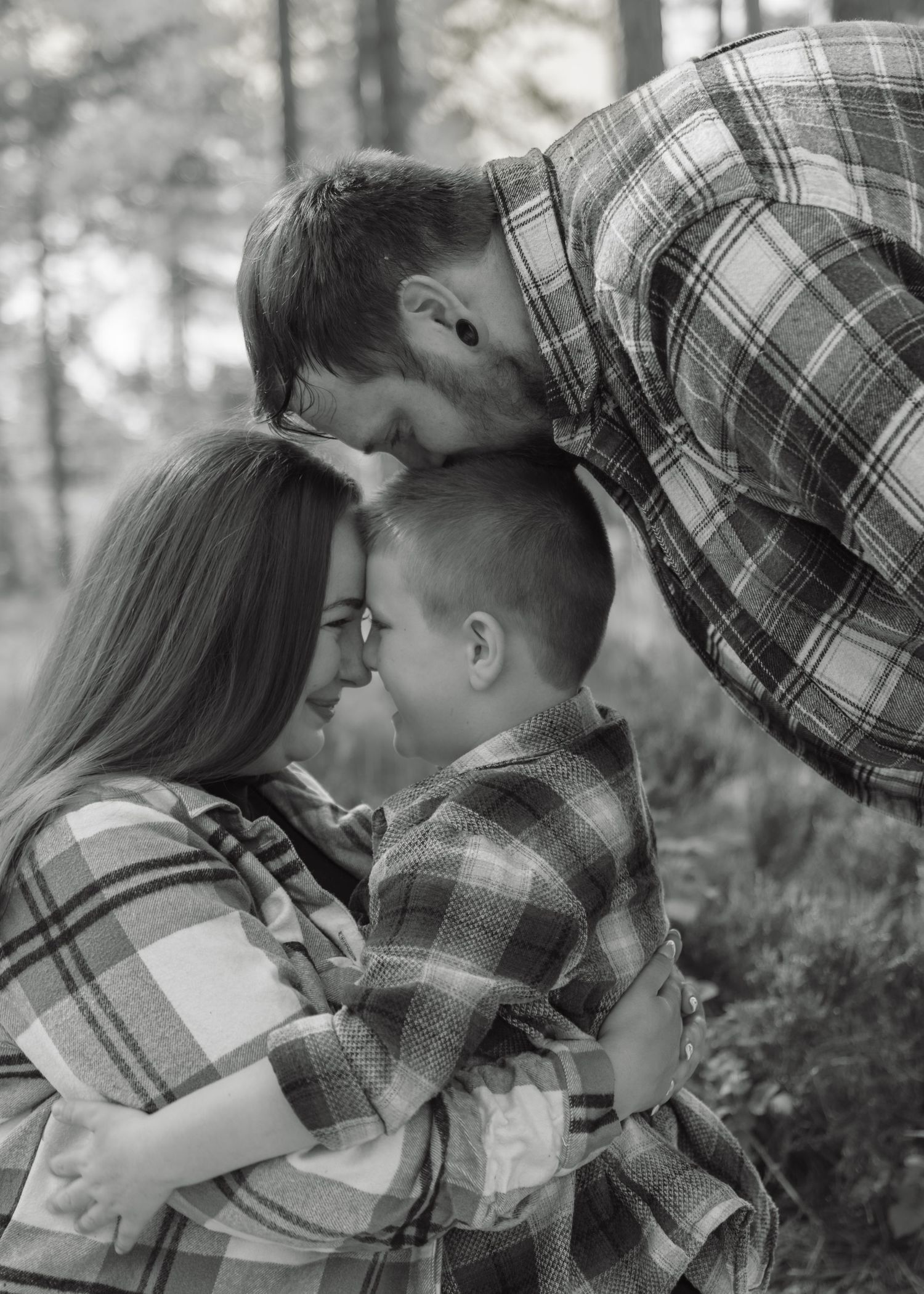 Black and white family photo of parents embracing child in plaid shirts outdoors in a forest setting.