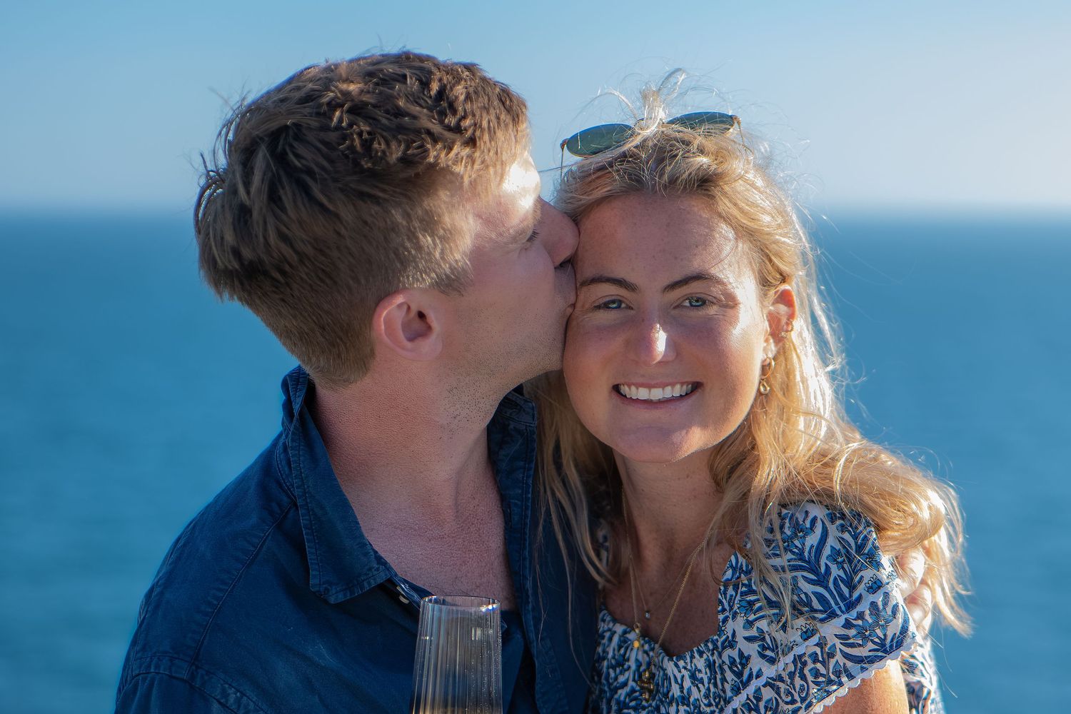Couple sharing a romantic kiss by the ocean, woman smiling holding champagne on a sunny day.