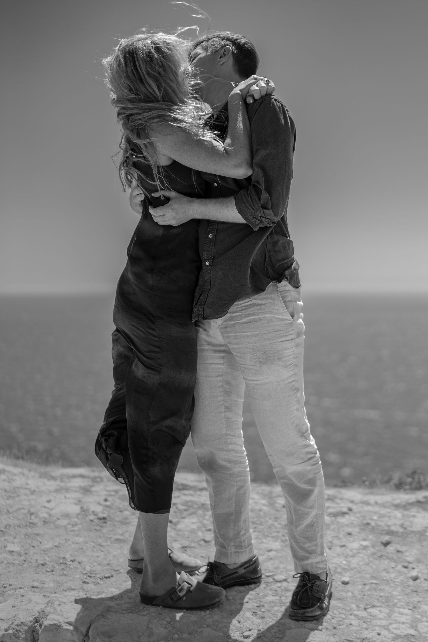 Romantic couple kissing and embracing on coastal cliff with ocean view in black and white photo.