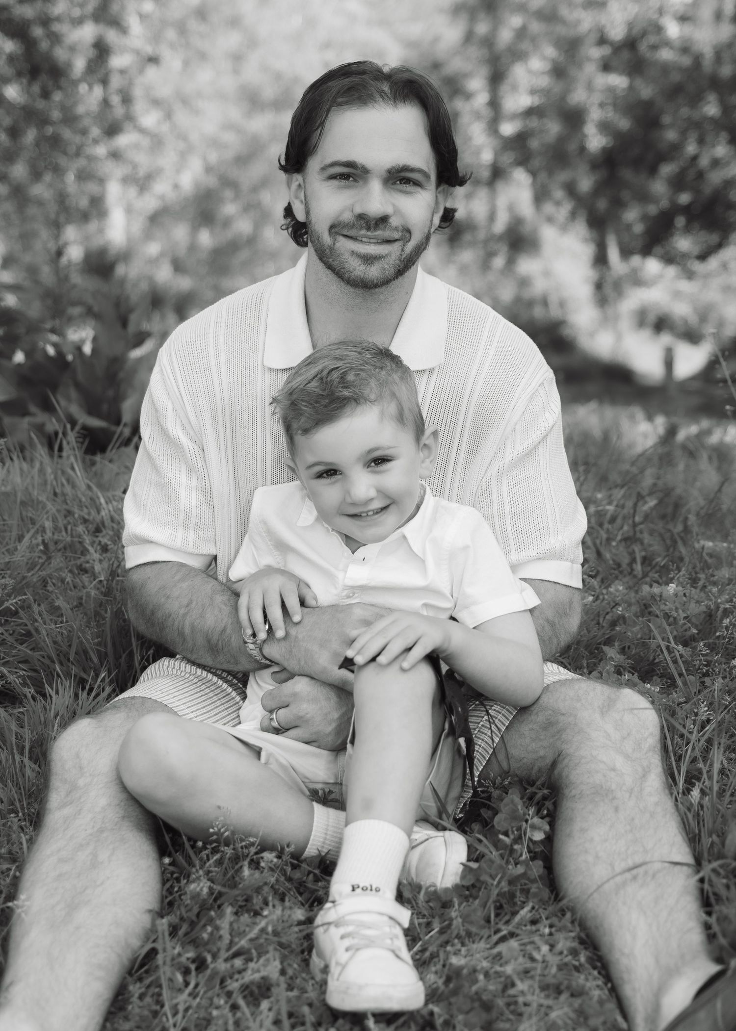 Black and white photo of a smiling man sitting on grass holding a young boy in a sunny outdoor setting.