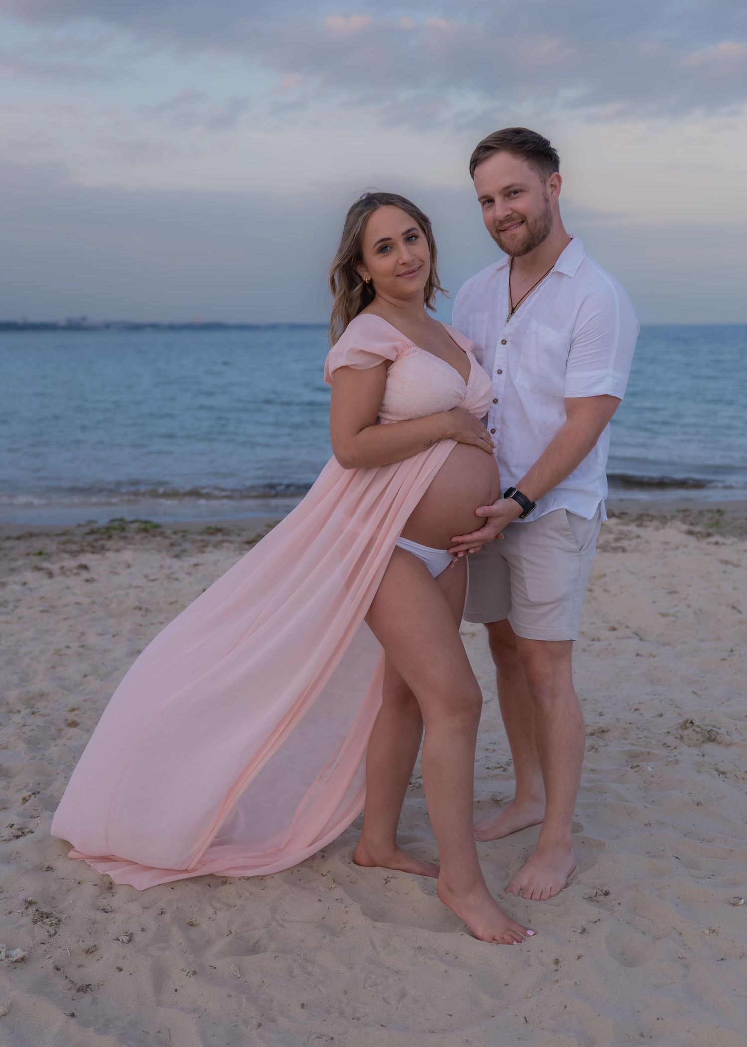 Pregnant couple posing on sandy beach at sunset, woman in pink maternity gown cradling baby bump.