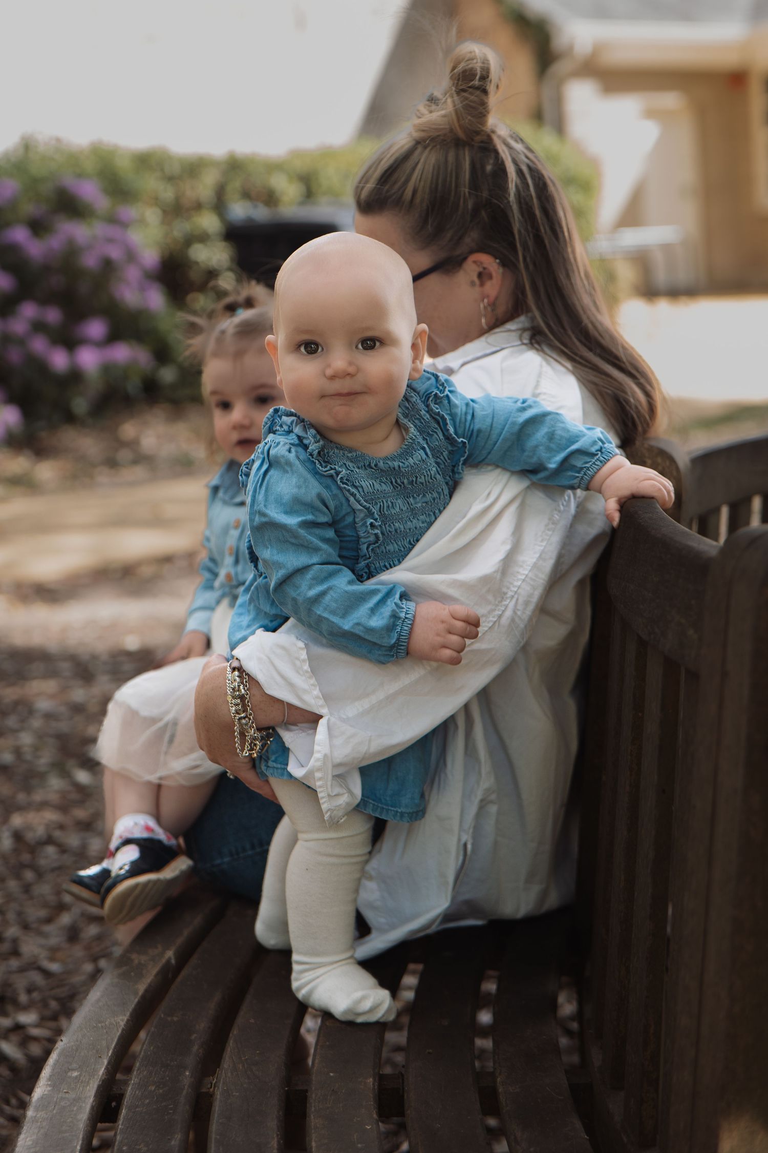 A mother sits on a park bench while her toddler daughter hugs a baby dressed in a blue denim outfit.