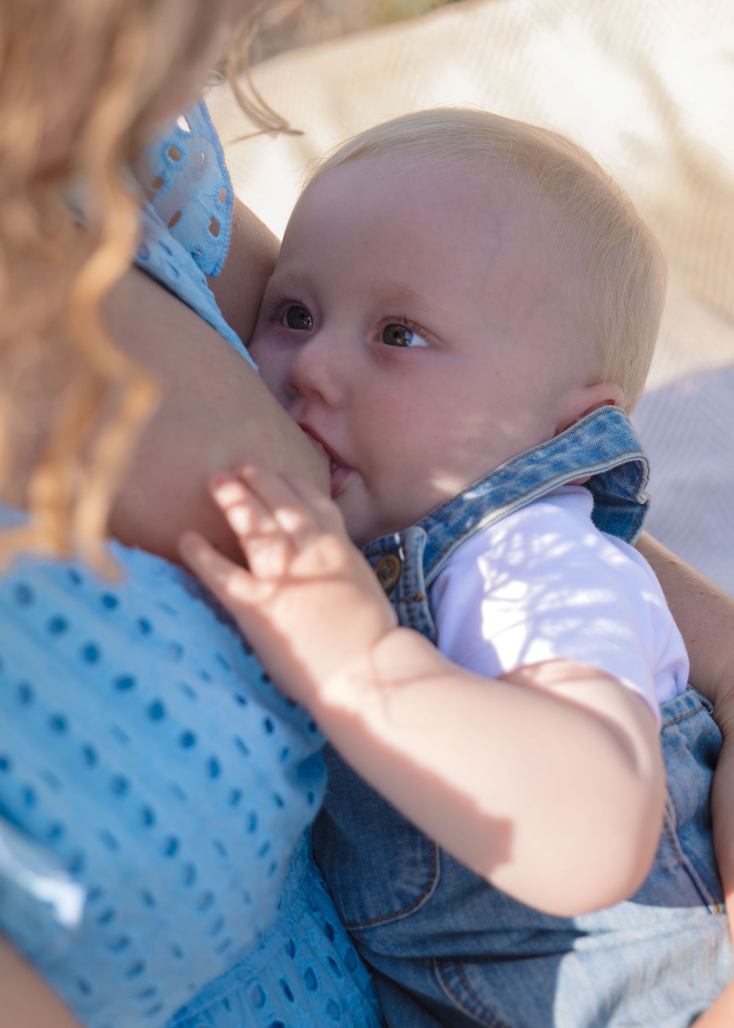 A baby in denim overalls breastfeeding outdoors in natural sunlight.