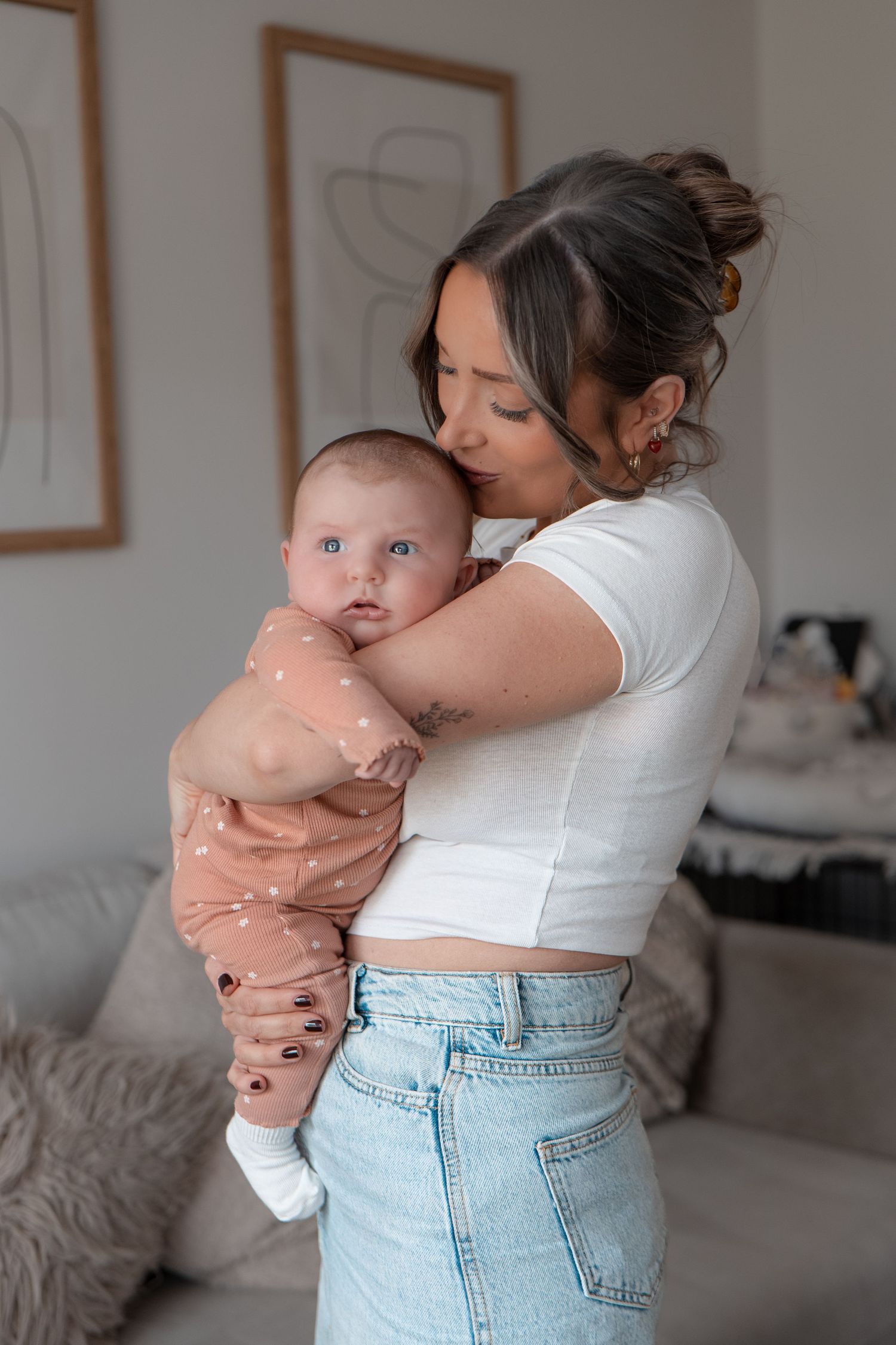 A young mother tenderly holds her blue-eyed baby dressed in a spotted peach outfit in a modern living room.