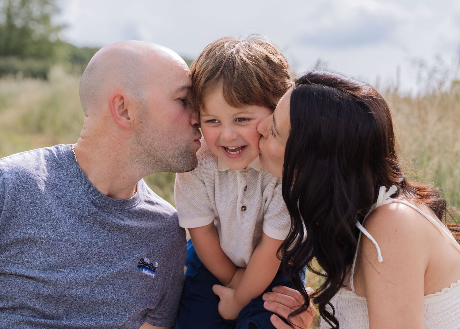 Parents kissing their laughing toddler boy on cheeks outdoors in a sunny field during a family photo session.