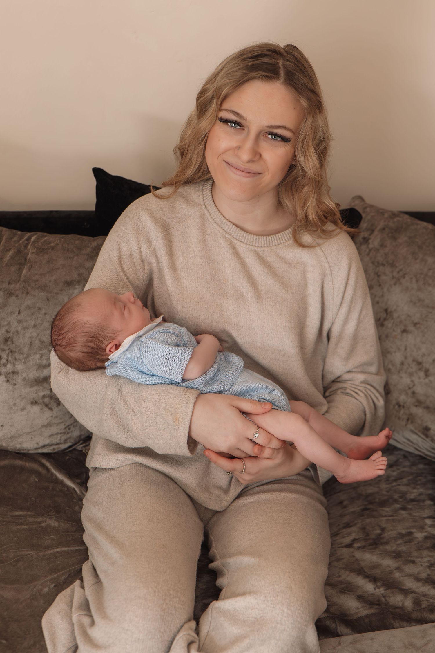 Smiling young mother sitting on a sofa holding her newborn baby in a blue outfit.