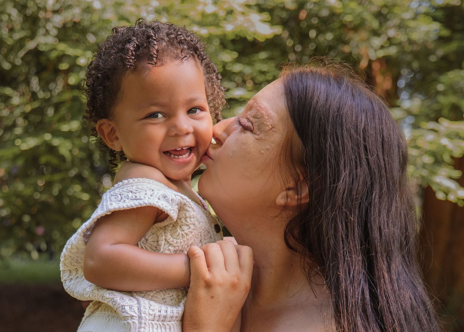 A mother lovingly kisses her smiling toddler on the cheek outdoors with lush green trees in the background.