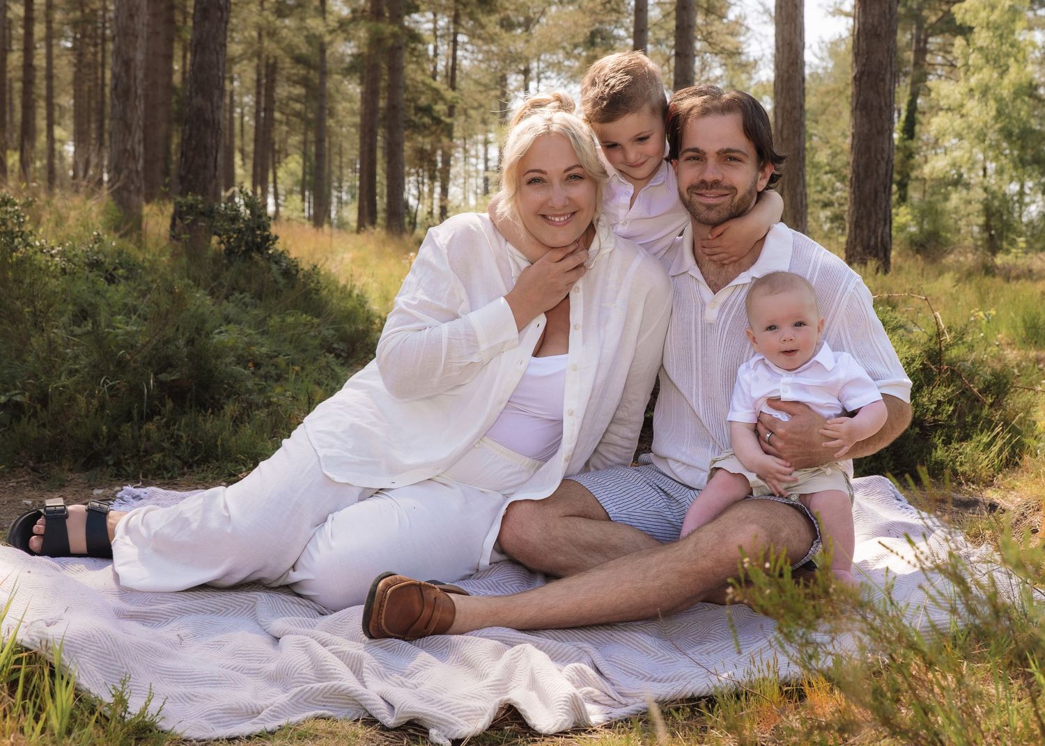 Happy family of four in white outfits sitting on a blanket in a forest setting for a professional photo.