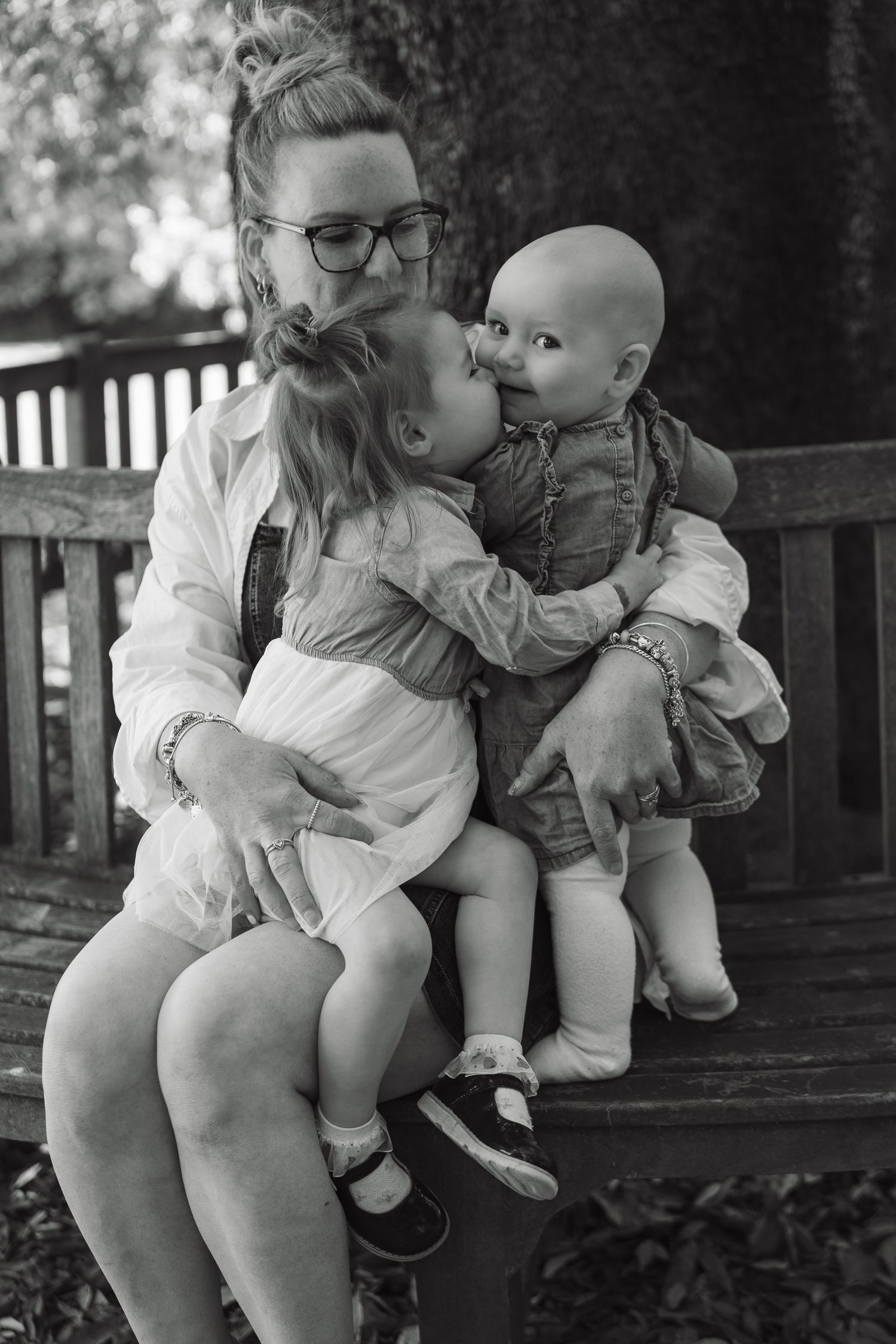 Black and white photo of a mother on a bench as her toddler daughter kisses her baby sibling tenderly.