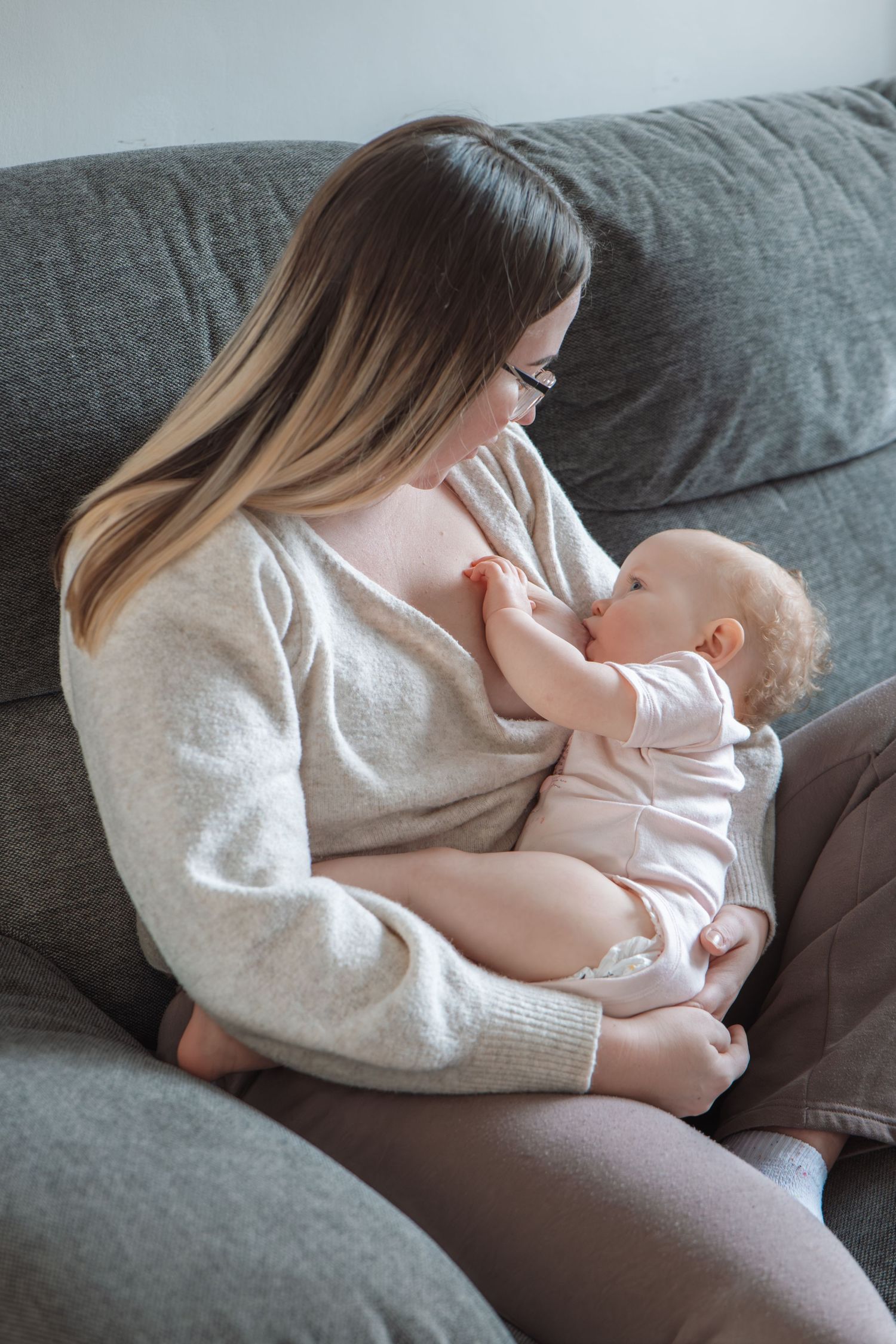 Young mother with glasses sitting on a gray sofa tenderly breastfeeding her baby girl.