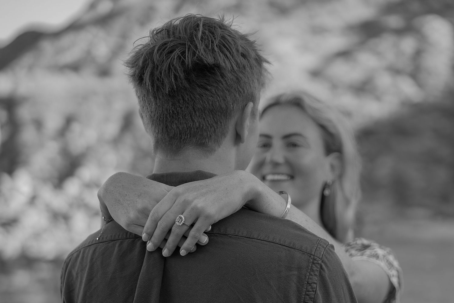 Black and white photo of a couple embracing outdoors, woman smiling with ring visible on hand resting on mans shoulder.