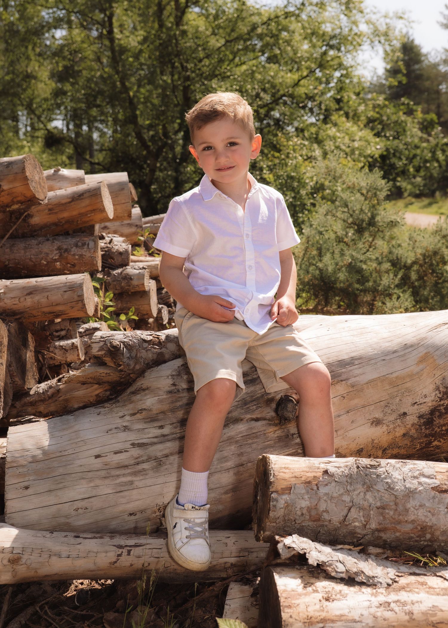 Young boy in white shirt and khaki shorts sitting on stacked logs outdoors in sunny woodland setting.