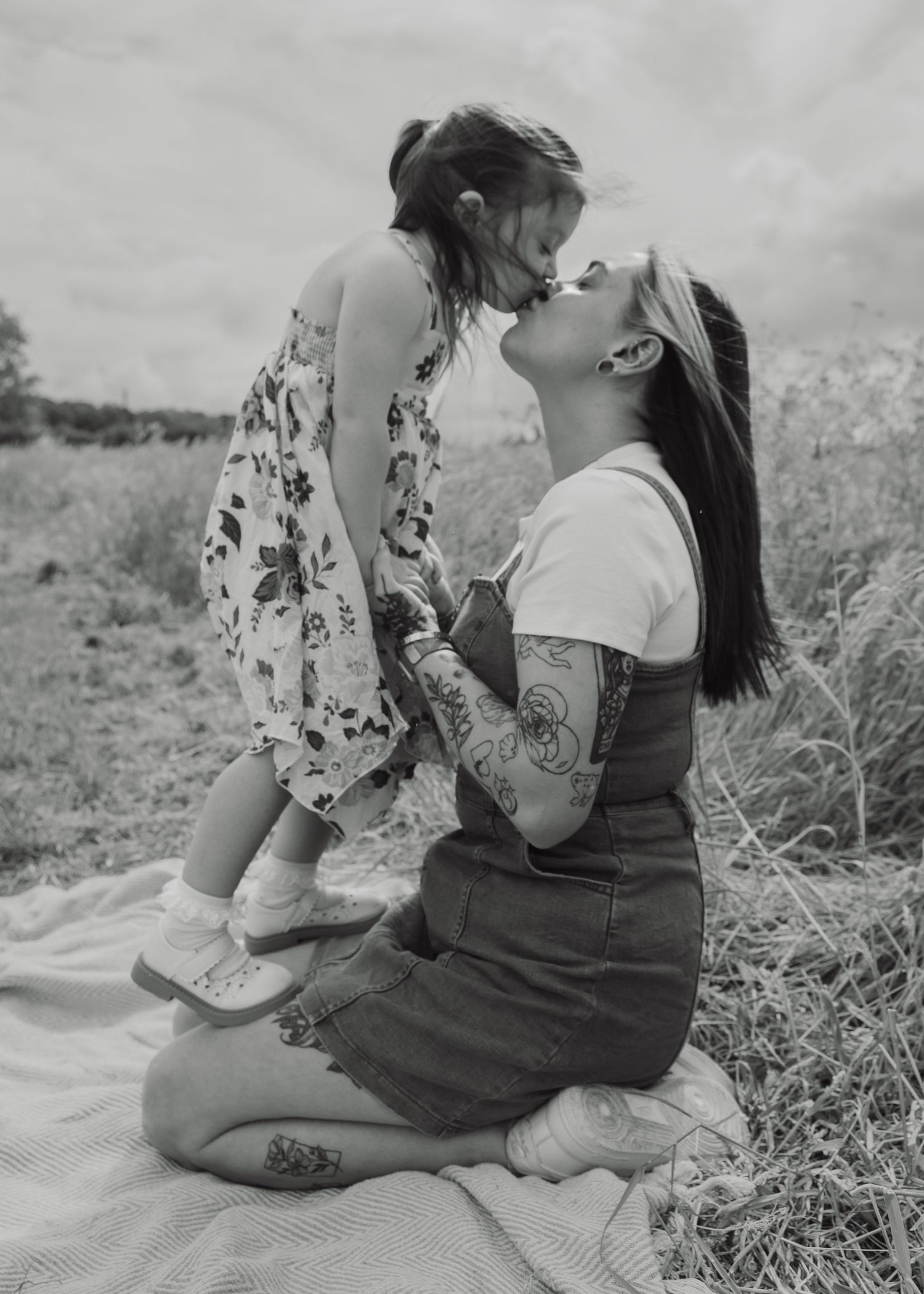 Black and white photo of tattooed mother kneeling in field, lovingly lifting and kissing her young daughter outdoors.