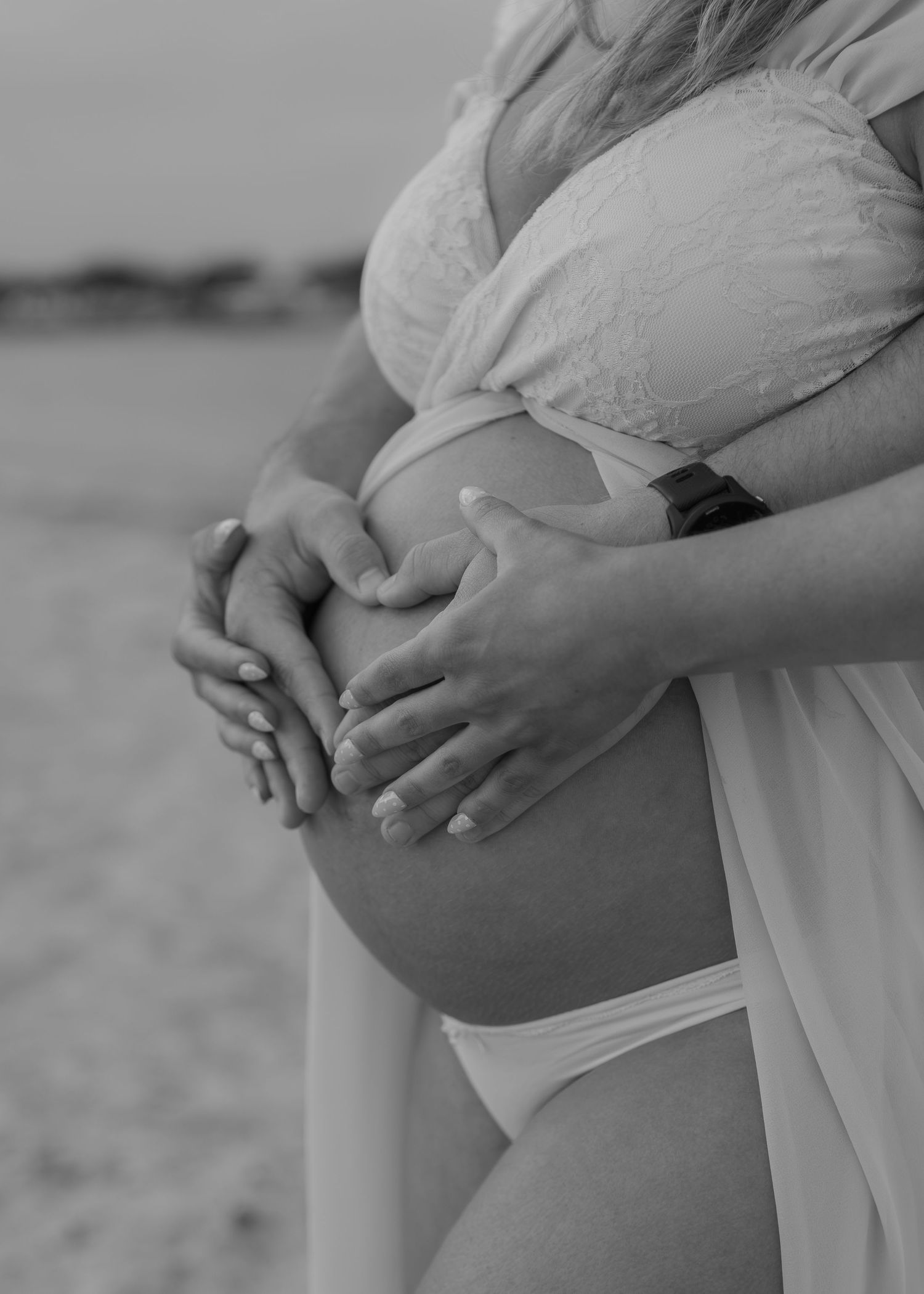 Black and white photo of a pregnant woman and partner forming a heart shape with their hands on her baby bump outdoors.