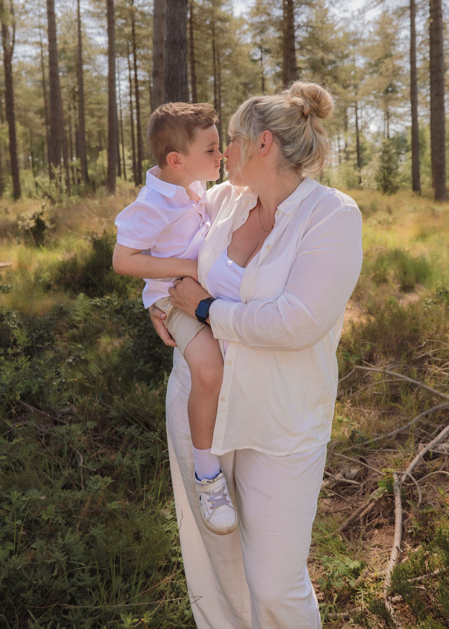 A mother in white holds her young son for a tender nose-to-nose moment in a sunlit forest setting.