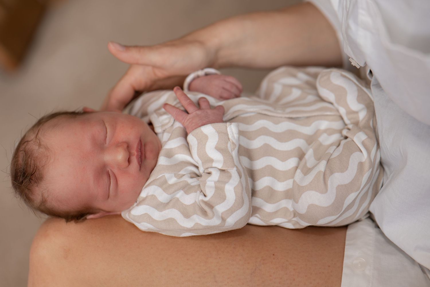 Newborn baby sleeping peacefully in a chevron print onesie, cradled in an adult's arms.