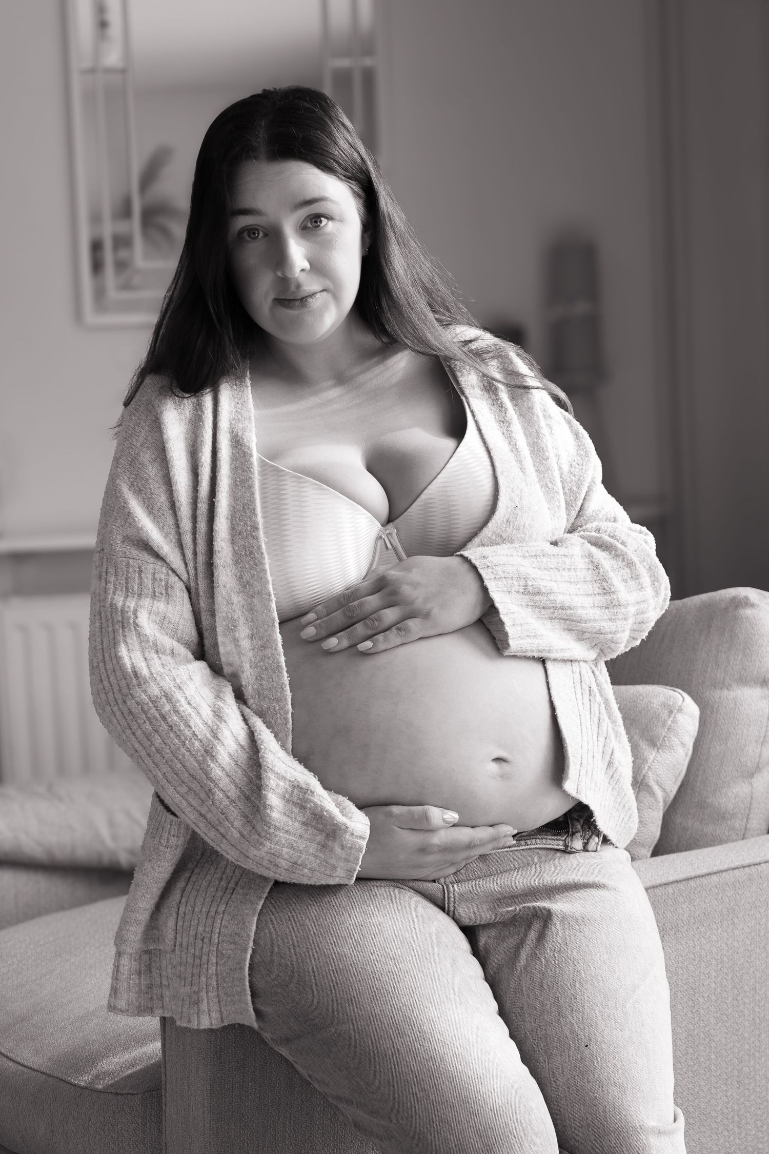 Black and white portrait of pregnant woman in bra and cardigan cradling her baby bump indoors.