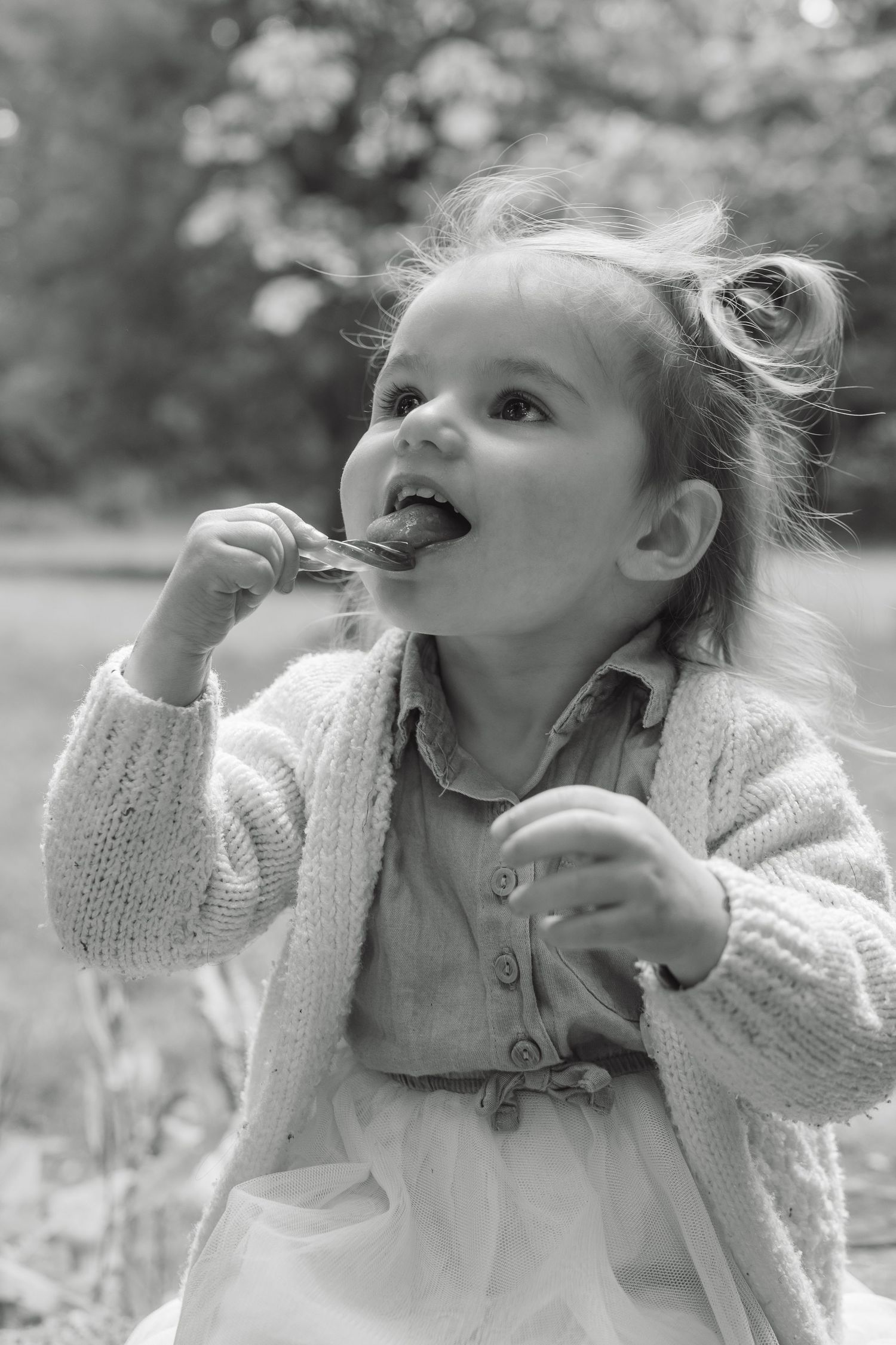 Black and white photo of a cute toddler girl eating a snack outdoors, wearing a knit cardigan.