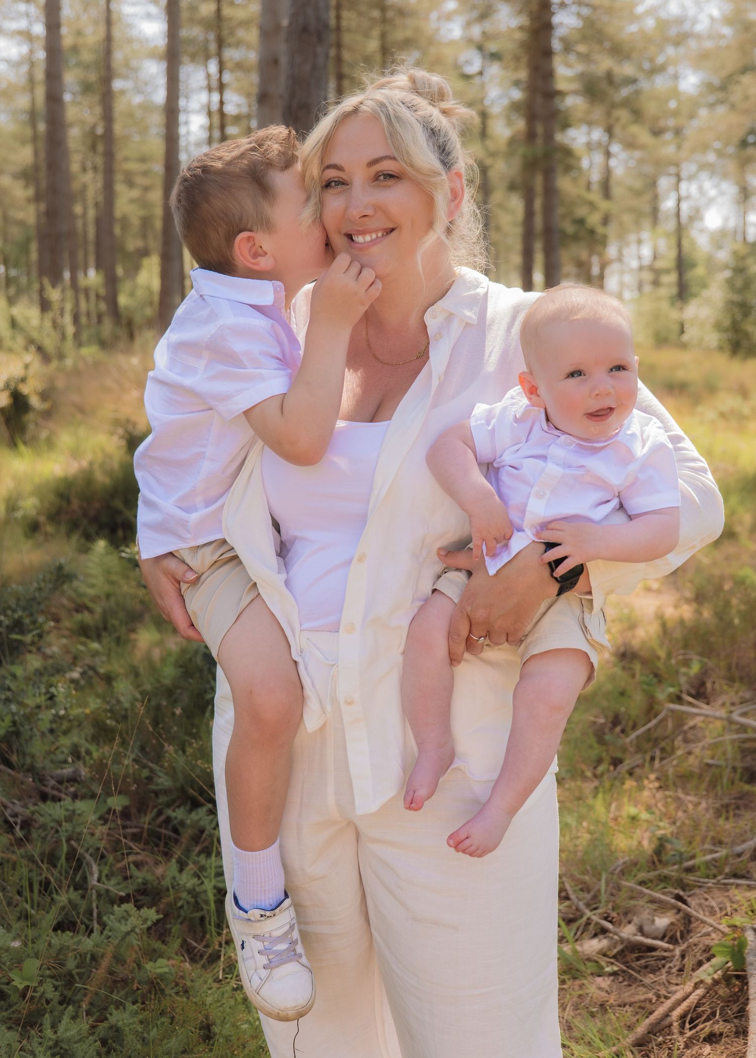 A smiling blonde woman holds two young boys in white shirts in a sunny forest setting.
