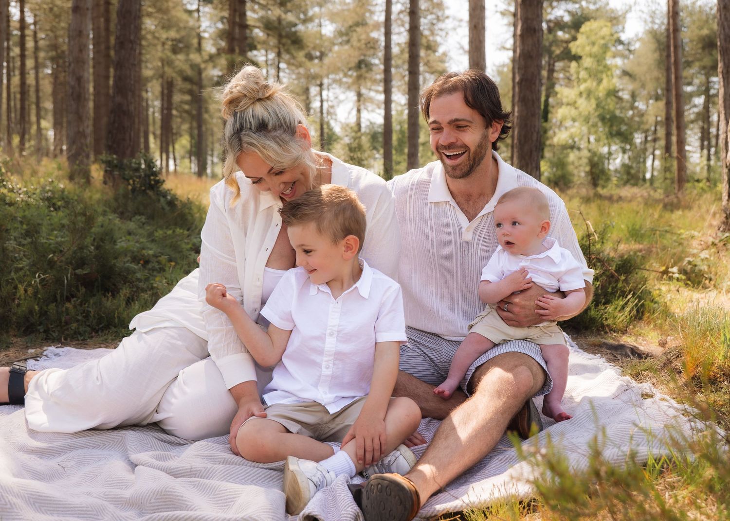 Happy family with two young children sitting on a blanket in a sunny pine forest for an outdoor picnic.