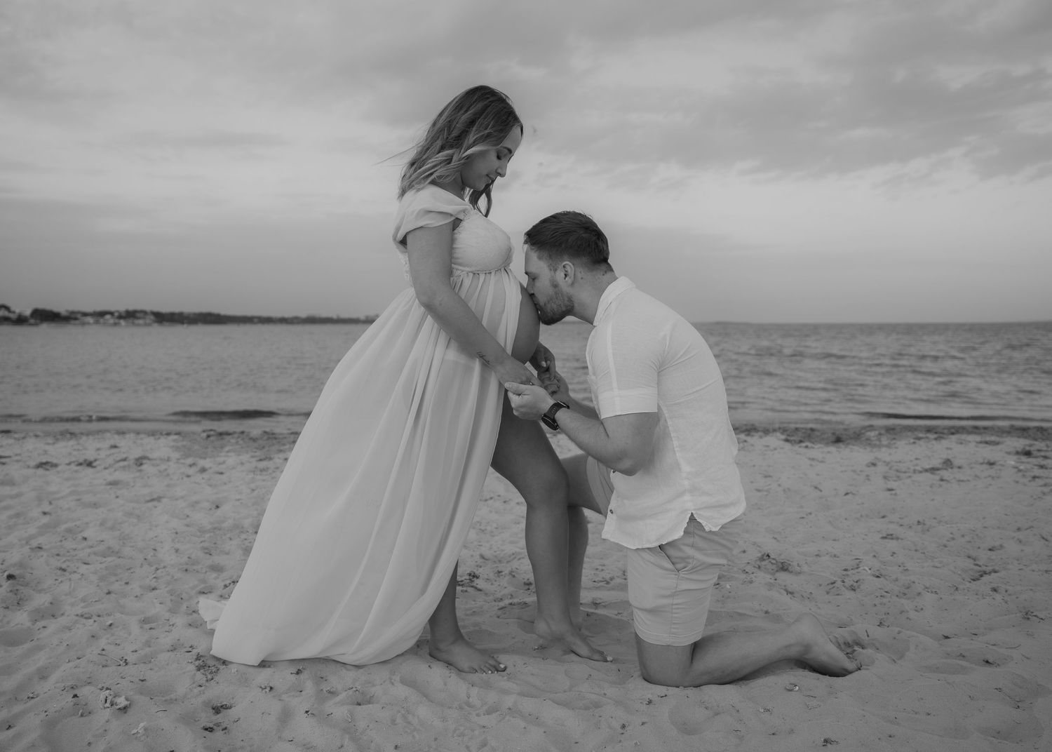 Black and white beach maternity photo of a man kneeling kissing pregnant womans belly on sandy shore.