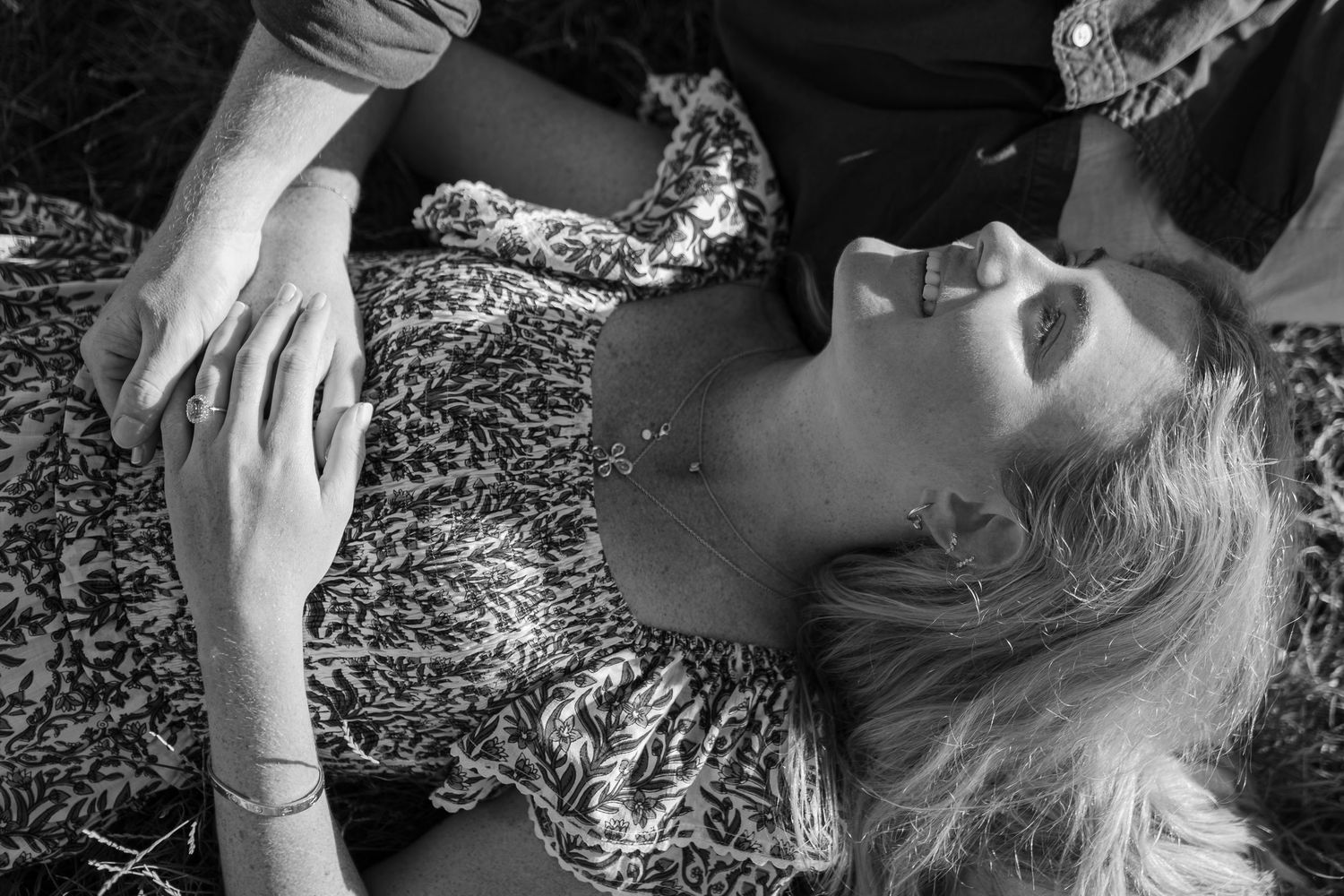 Black and white photo of a woman lying in grass, laughing, wearing a floral dress, with a man beside her.