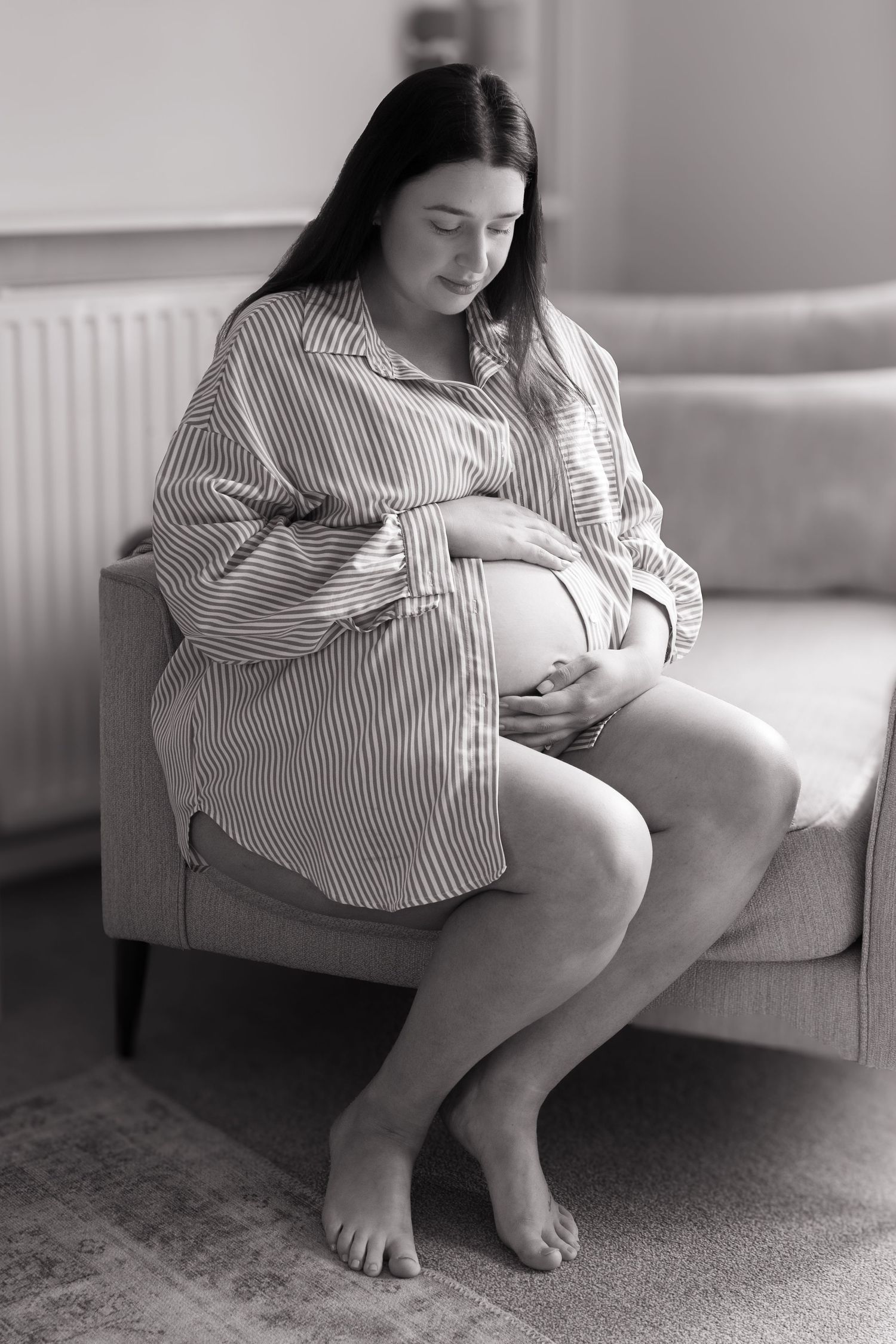 Black and white photo of a pregnant woman in a striped shirt sitting on a sofa, cradling her baby bump.