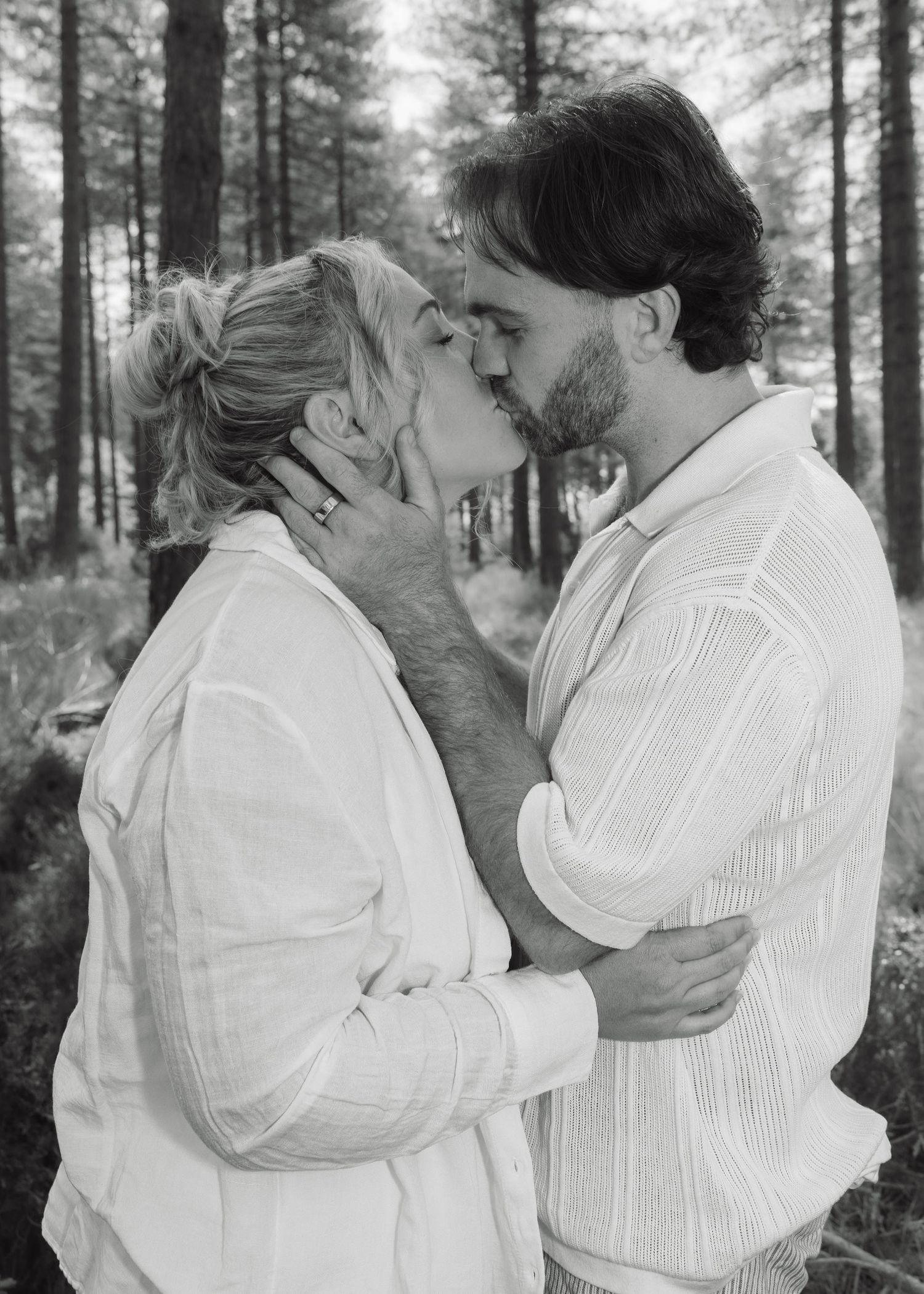 A couple shares a romantic kiss in a black and white forest photo, both wearing white shirts.