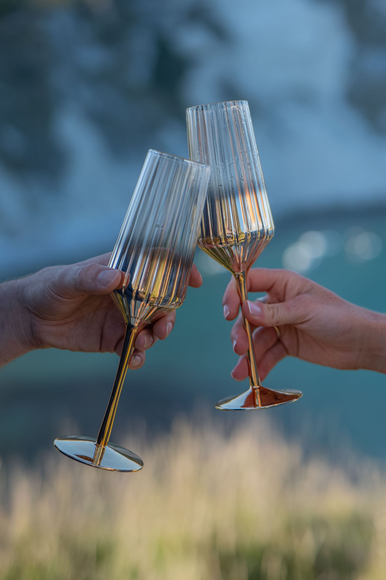 Two hands clinking elegant gold-stemmed ribbed champagne flutes outdoors in a celebratory toast.