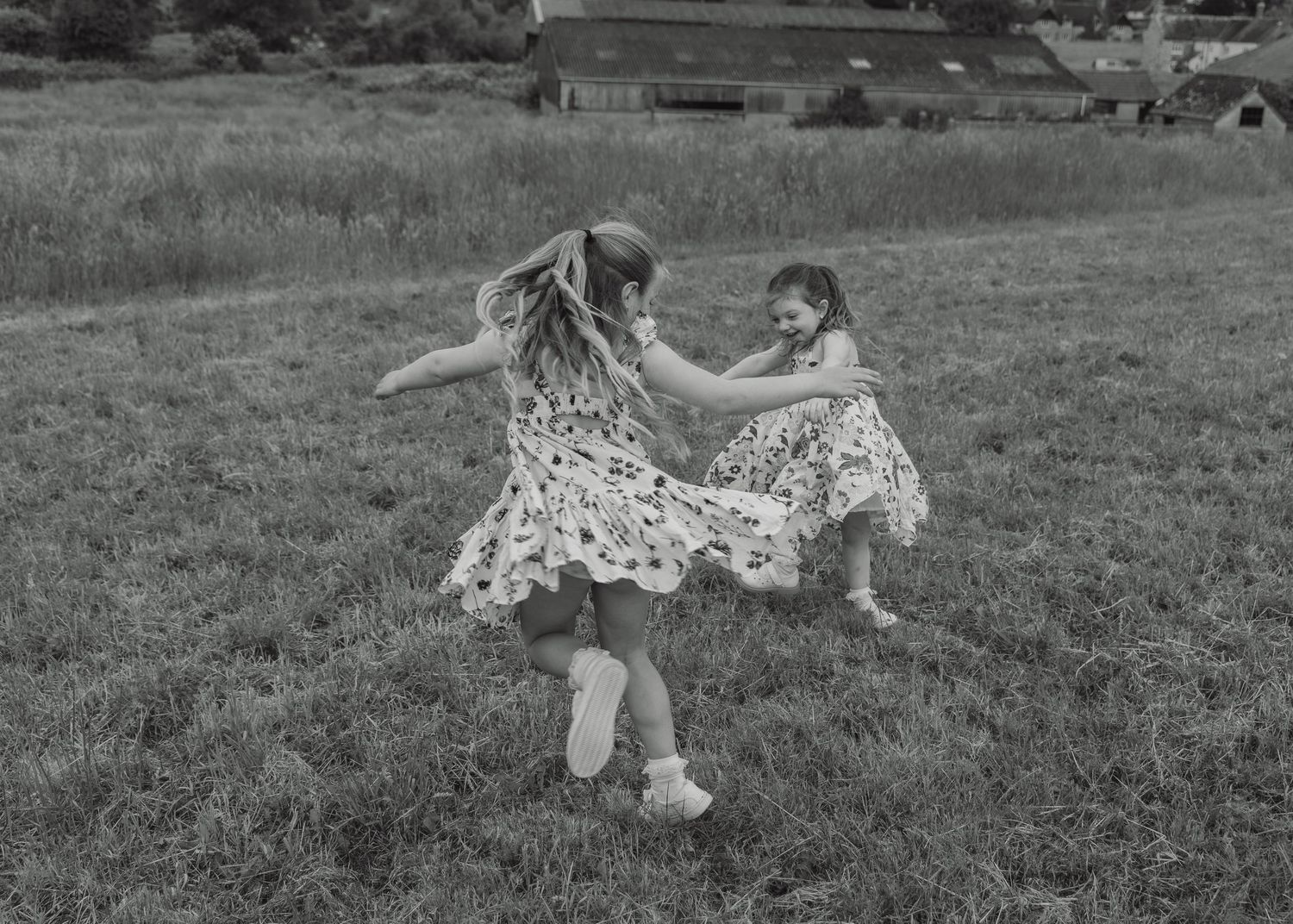 Two young girls in matching dresses spinning and playing together in a grassy field in black and white.