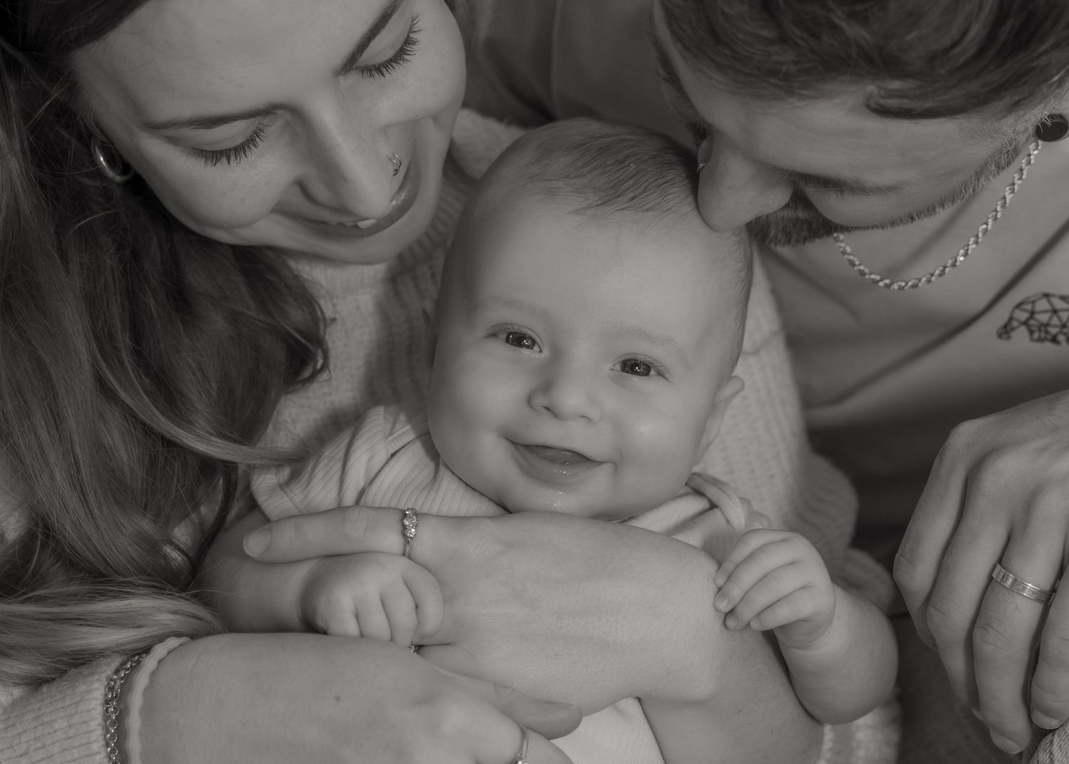Black and white photo of two parents smiling and cuddling their happy, smiling newborn baby.
