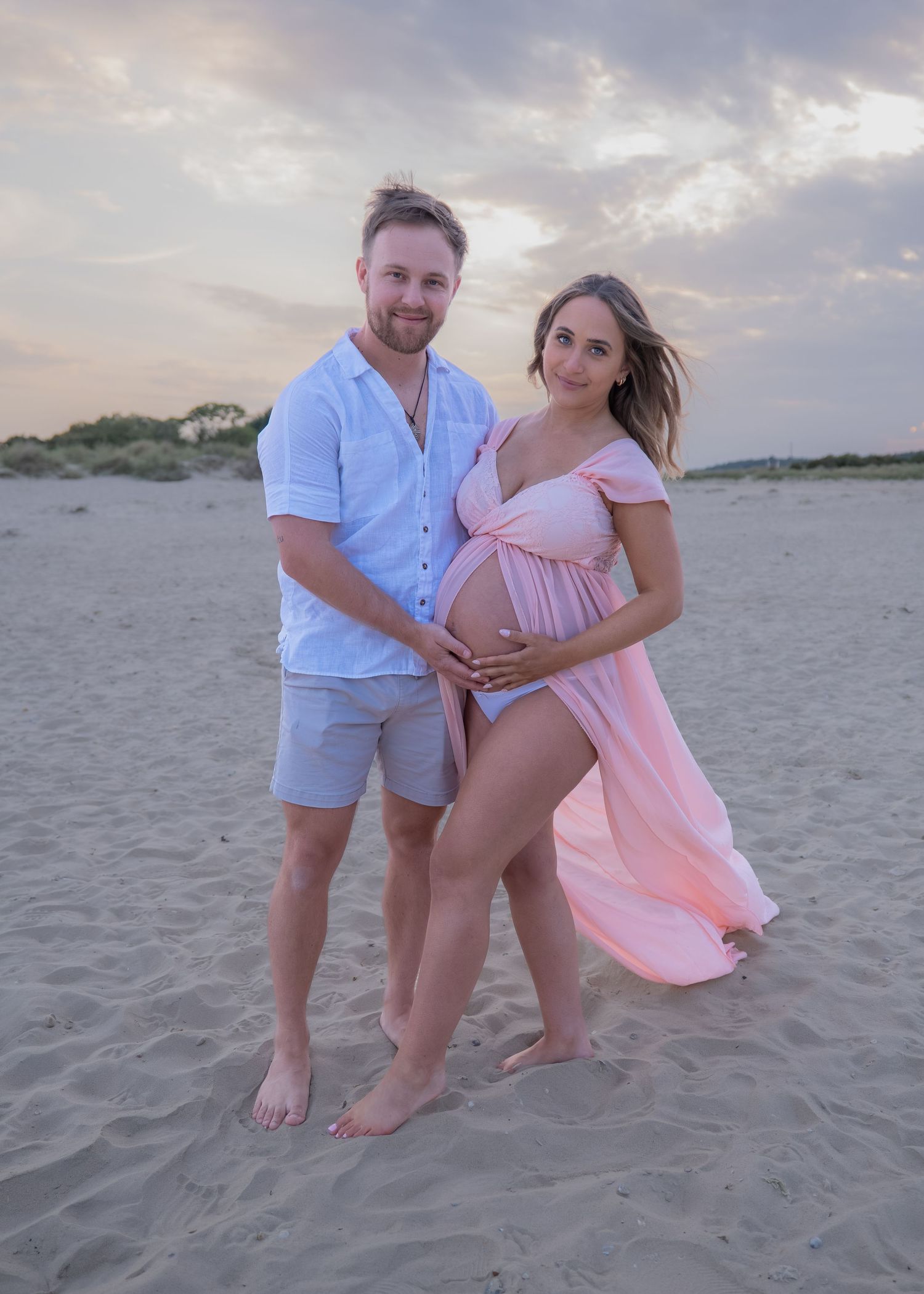 Couple poses on sandy beach at sunset, pregnant woman in pink maternity gown, man in light blue shirt and shorts.