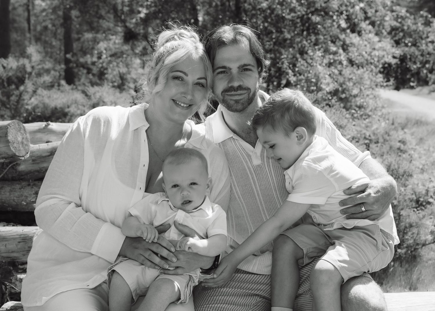 Black and white family portrait of smiling parents with two young children seated outdoors among trees.