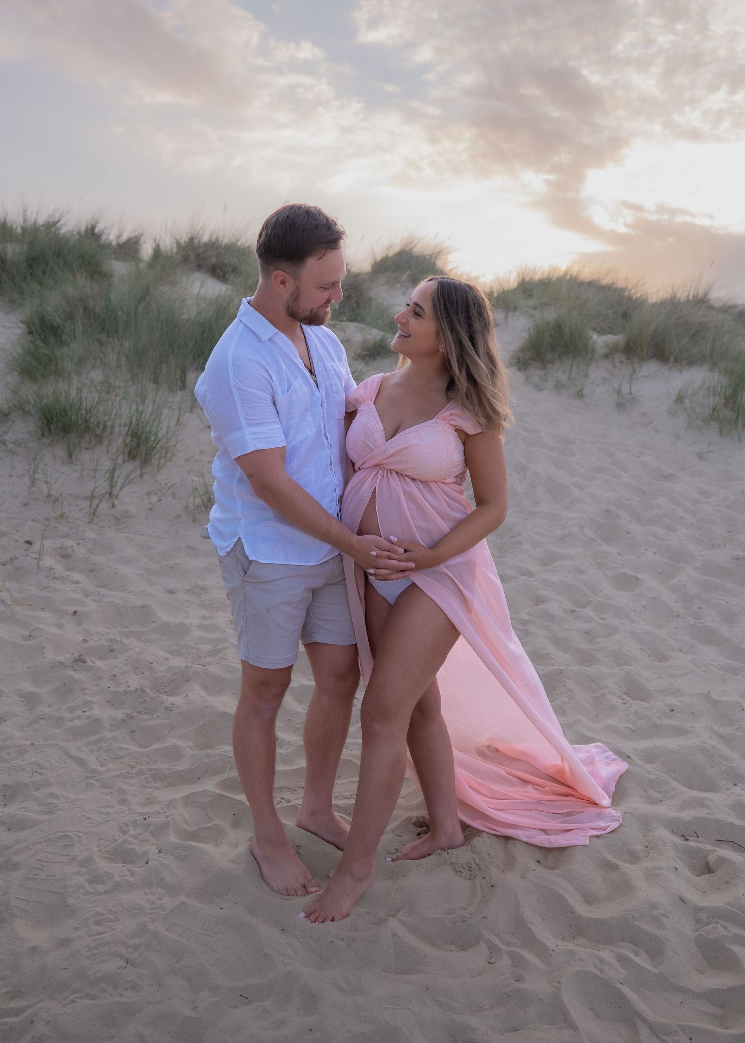 Couple in maternity photoshoot on sandy beach at sunset, woman in pink flowing gown cradling baby bump.