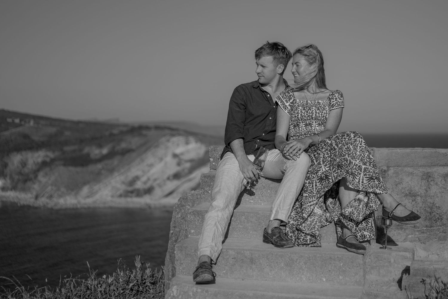 Black and white photo of a romantic couple sitting together on a stone wall with coastal cliffs in the background.