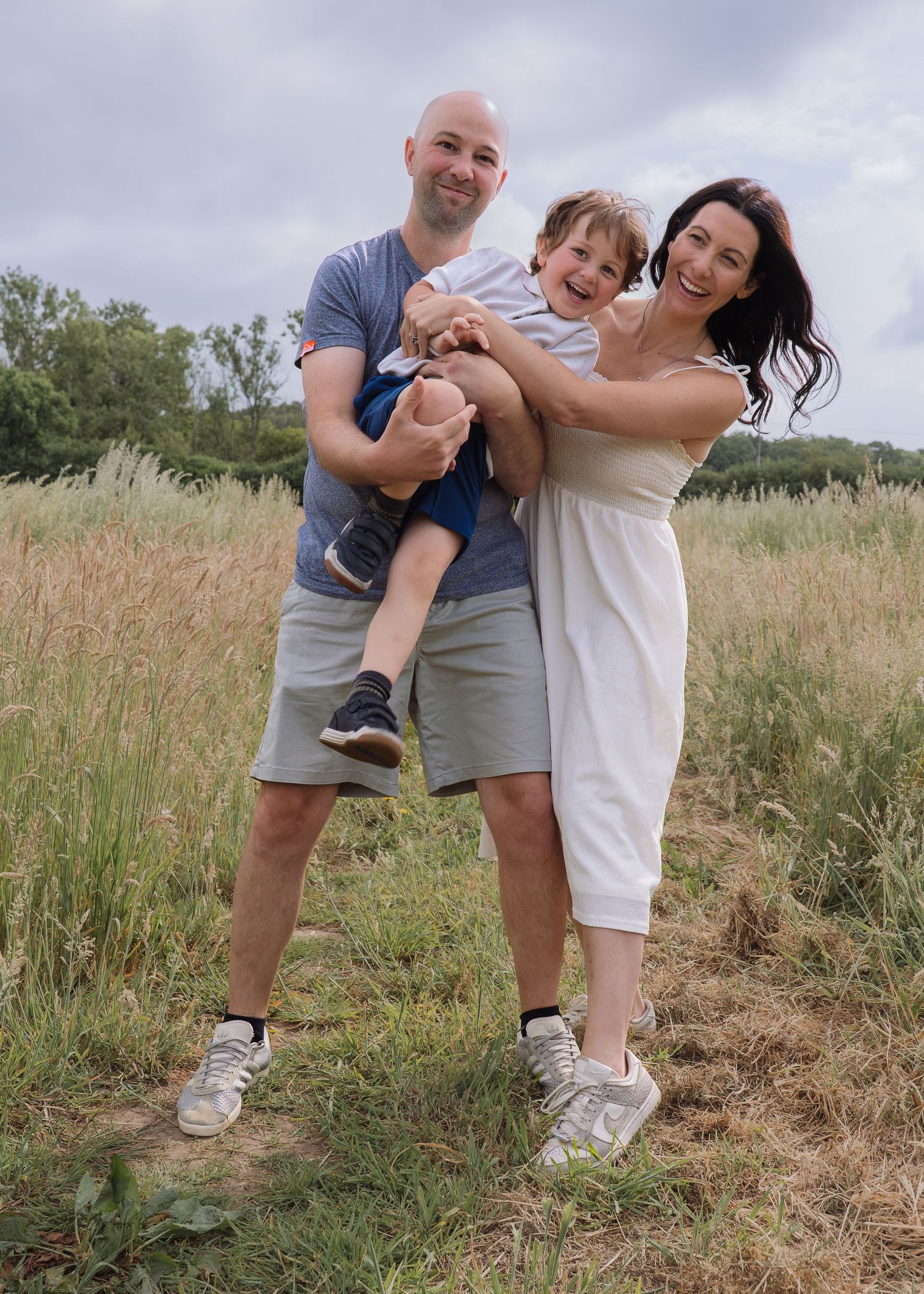 A happy family of three poses in a sunny meadow, with a bald man and dark-haired woman holding a laughing child.