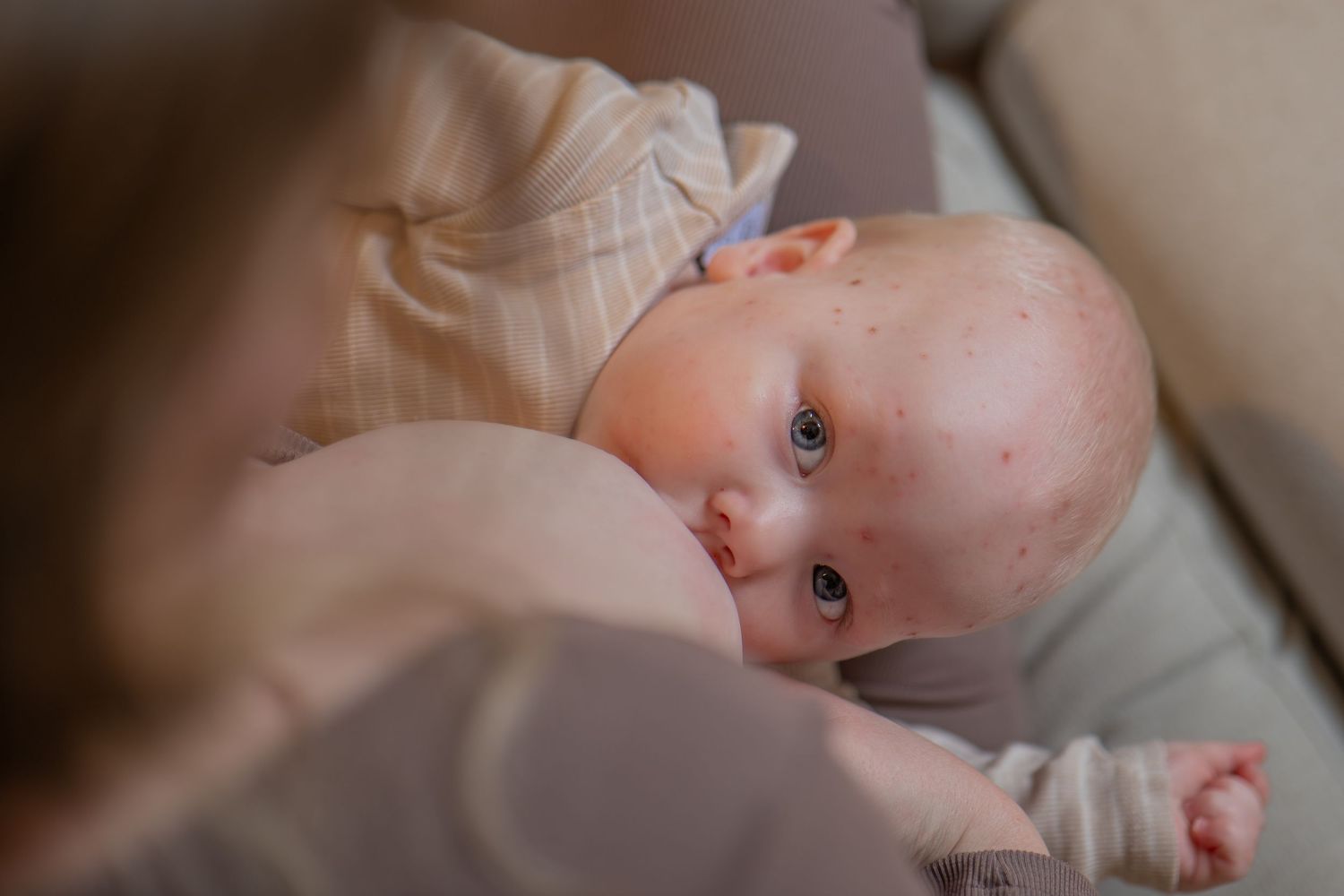 Newborn baby with blue eyes breastfeeding, cradled in mother's arms on a soft couch.