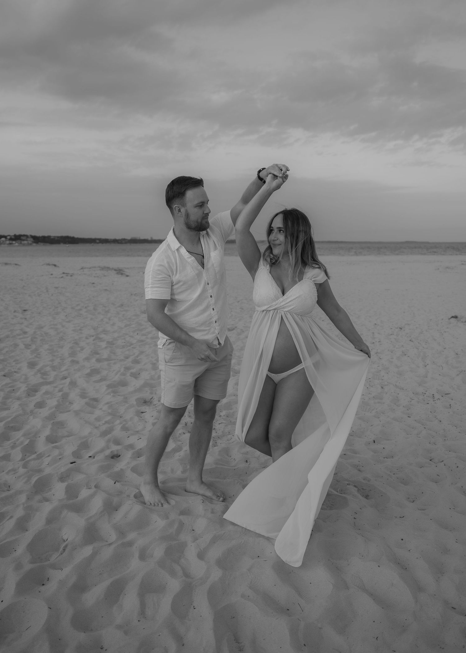 Black and white photo of a couple dancing on a beach, woman in flowing white dress being twirled at sunset.
