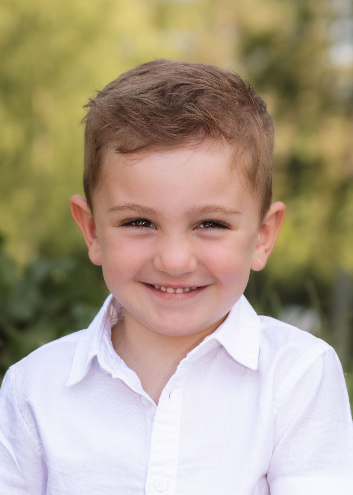 A smiling young boy with brown hair wearing a white collared shirt poses for a portrait outdoors.
