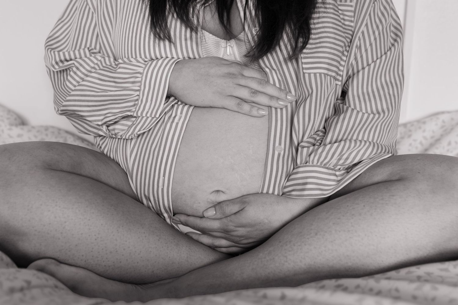 Black and white photo of pregnant woman in striped shirt sitting cross-legged holding baby bump.