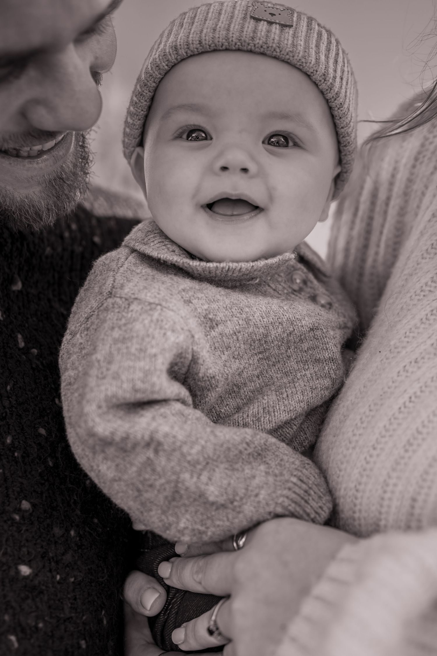 Smiling baby in knit hat and sweater held by parents in black and white photo.