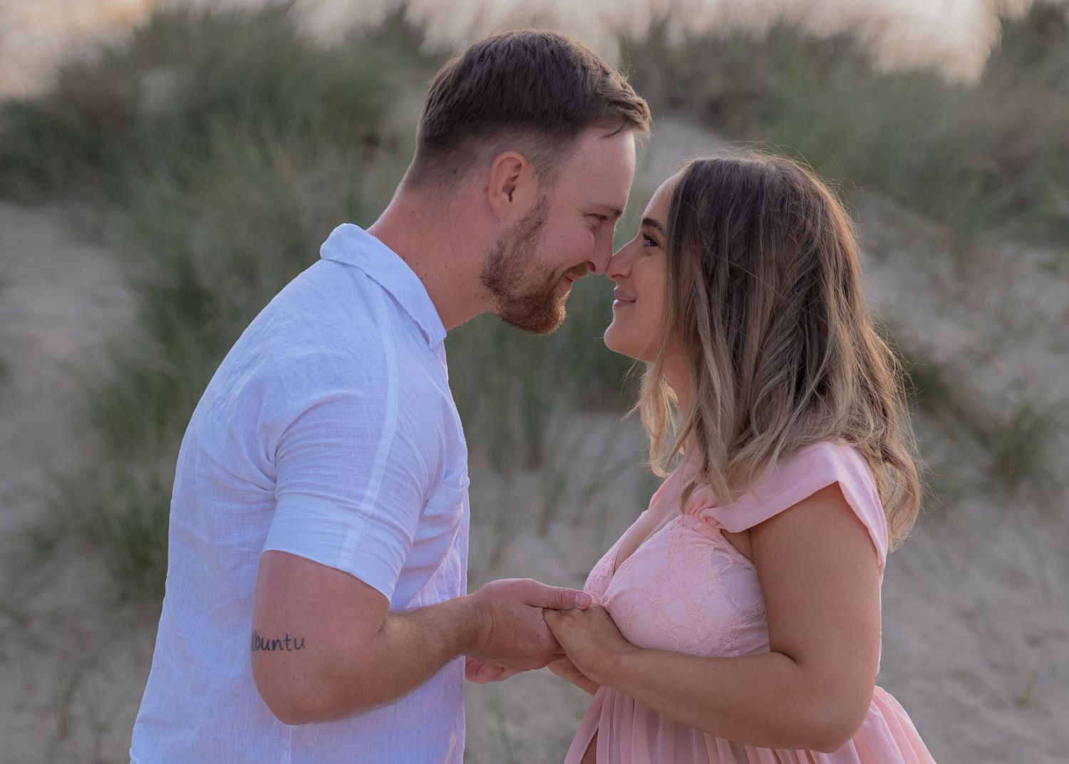 Couple sharing a romantic moment on a sandy beach, smiling and holding hands with sand dunes in the background.