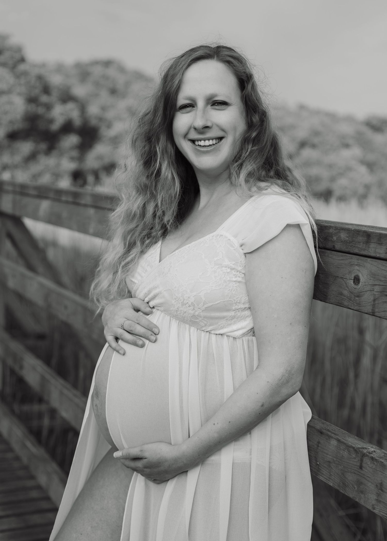 Black and white maternity photo of smiling pregnant woman in white dress holding belly on wooden bridge outdoors.