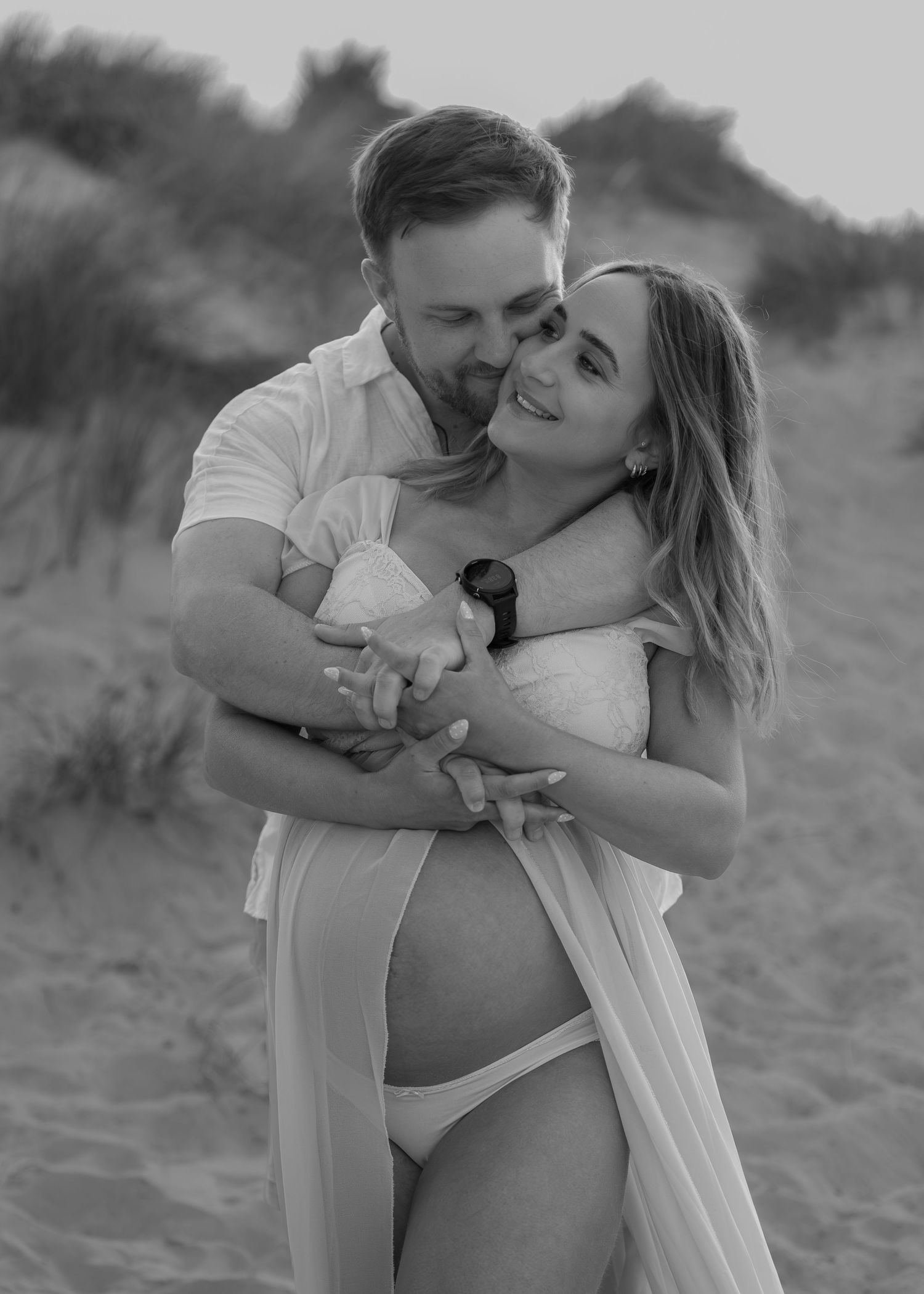 Black and white maternity photo of a couple embracing on a beach, the woman showing her baby bump in a white outfit.