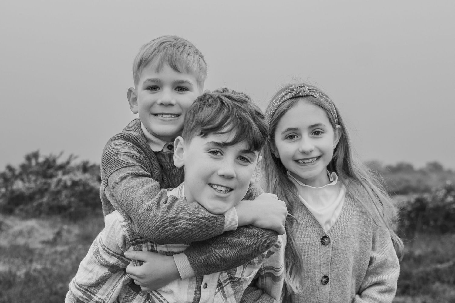 Black and white photo of three happy siblings hugging outdoors in a field.