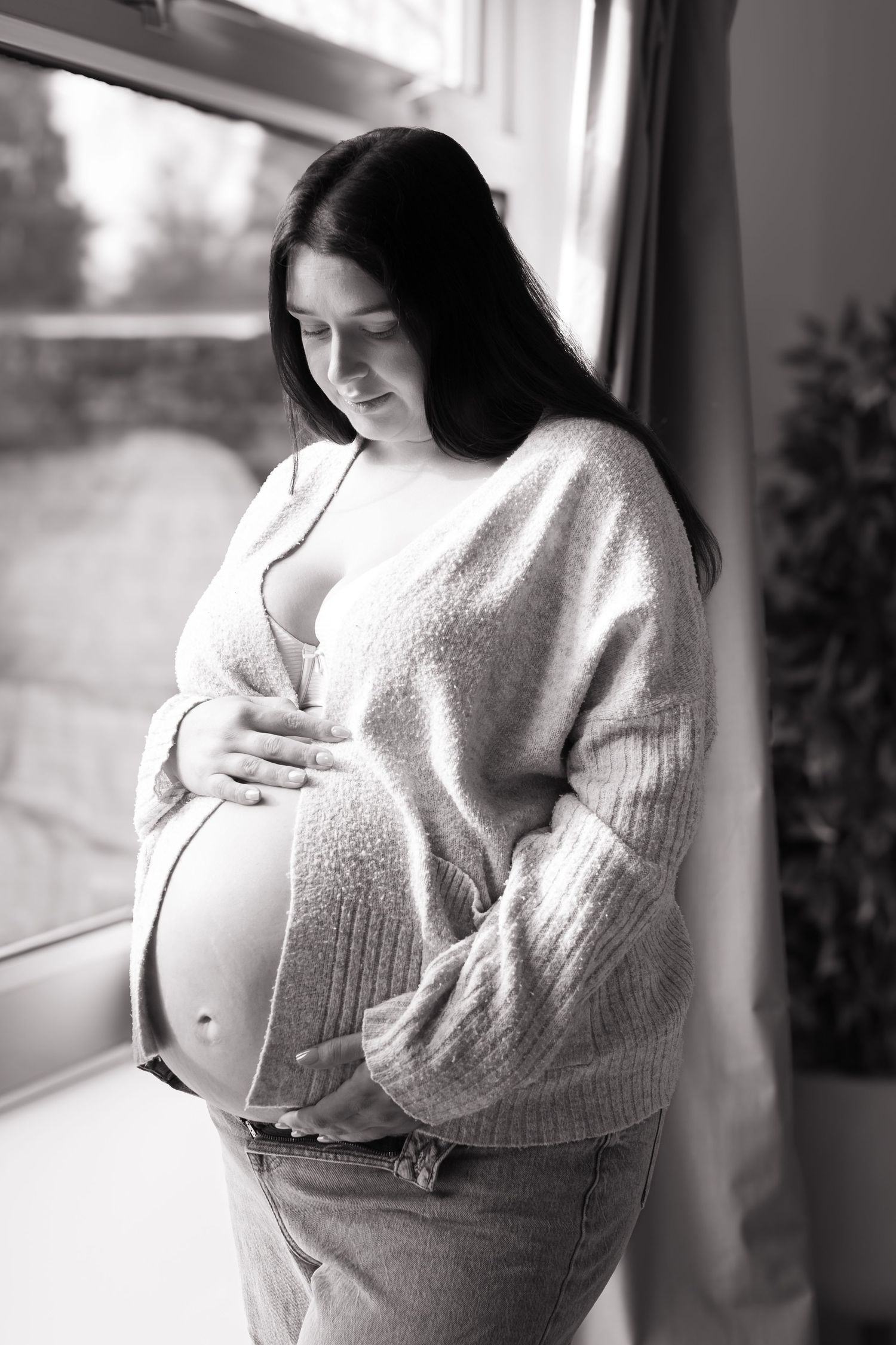 Black and white photo of pregnant woman in cardigan holding baby bump by a window.