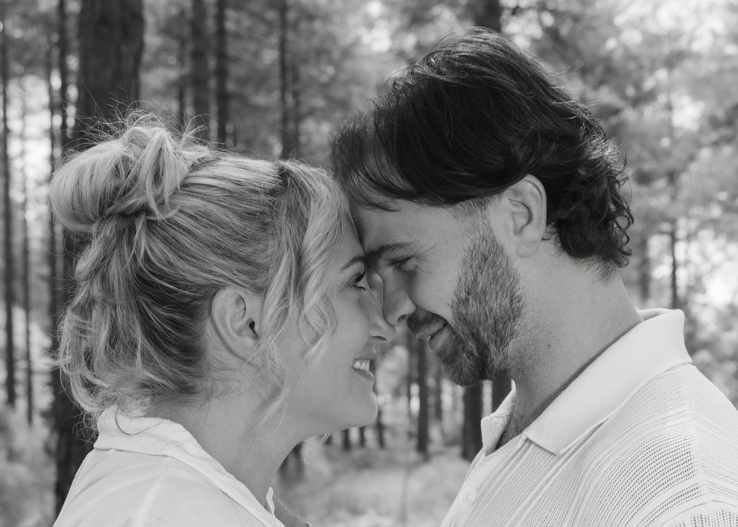 Black and white photo of a romantic couple smiling and leaning in close together outdoors among trees.