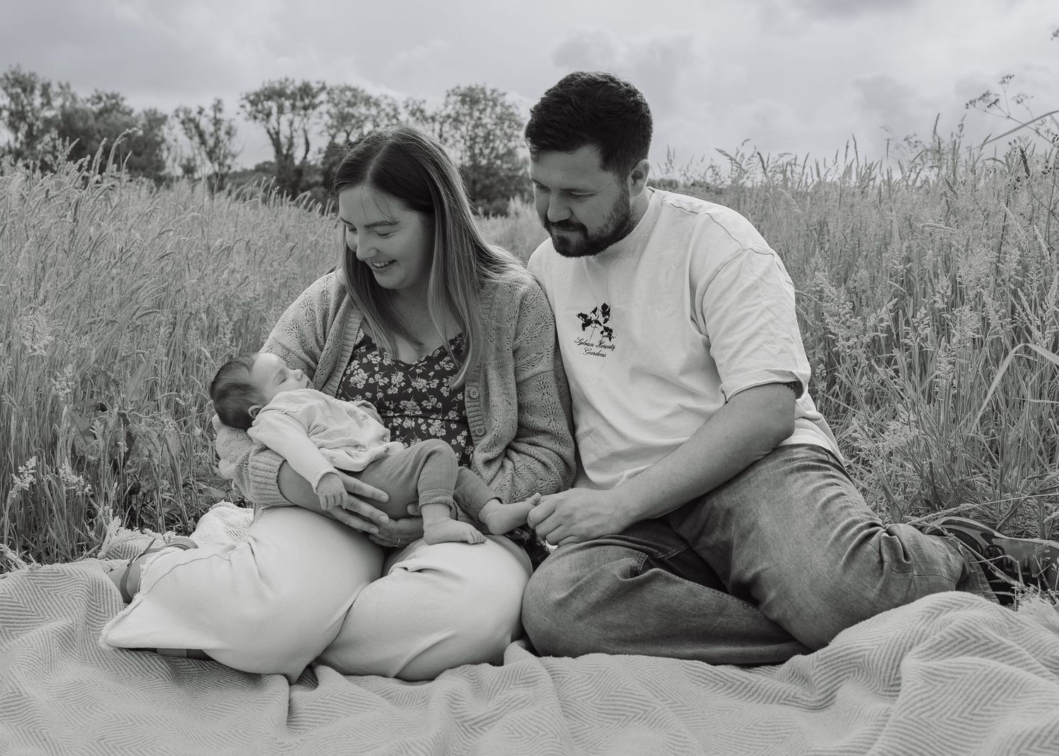 Black and white photo of a couple sitting in a field holding their newborn baby on a blanket outdoors.