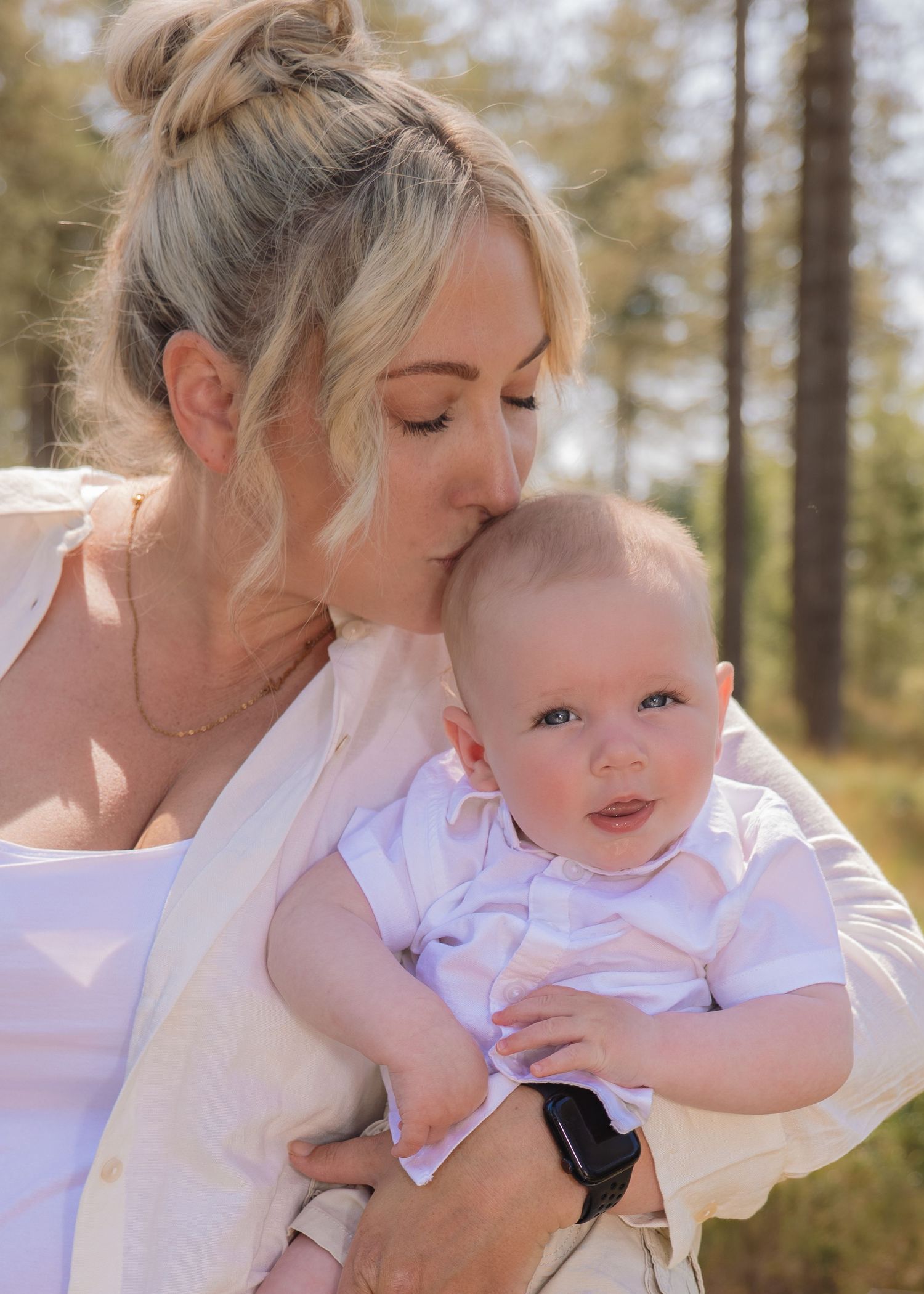 A blonde woman kisses a baby boy in white outdoors among trees on a sunny day.