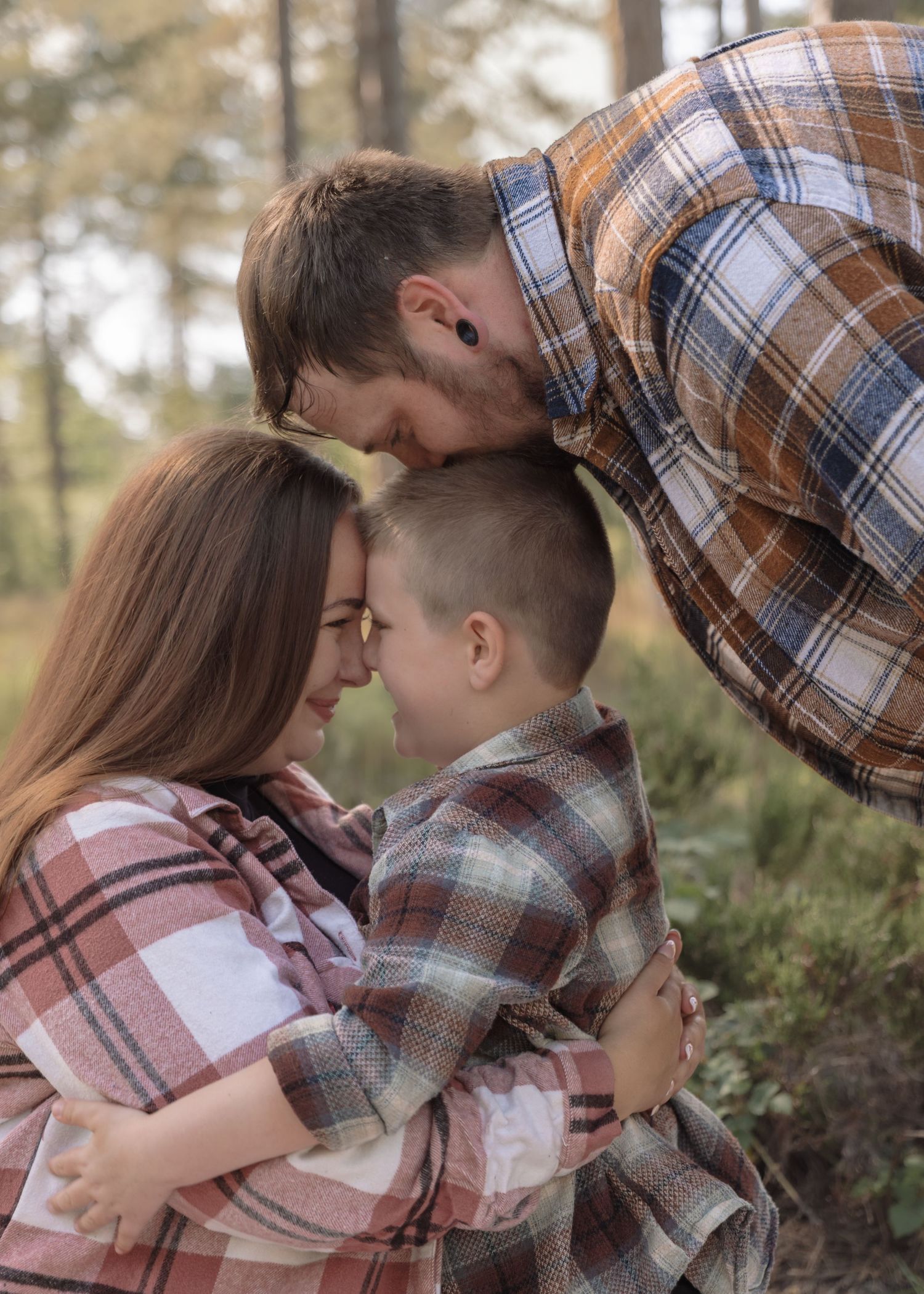 A family of three sharing a tender embrace outdoors, wearing coordinating plaid shirts in a forest setting.